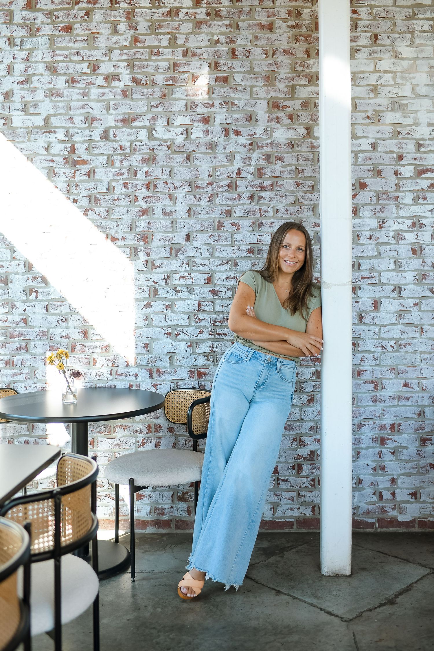 Woman in wide-leg jeans and green top leaning against white column in modern cafe with brick wall.