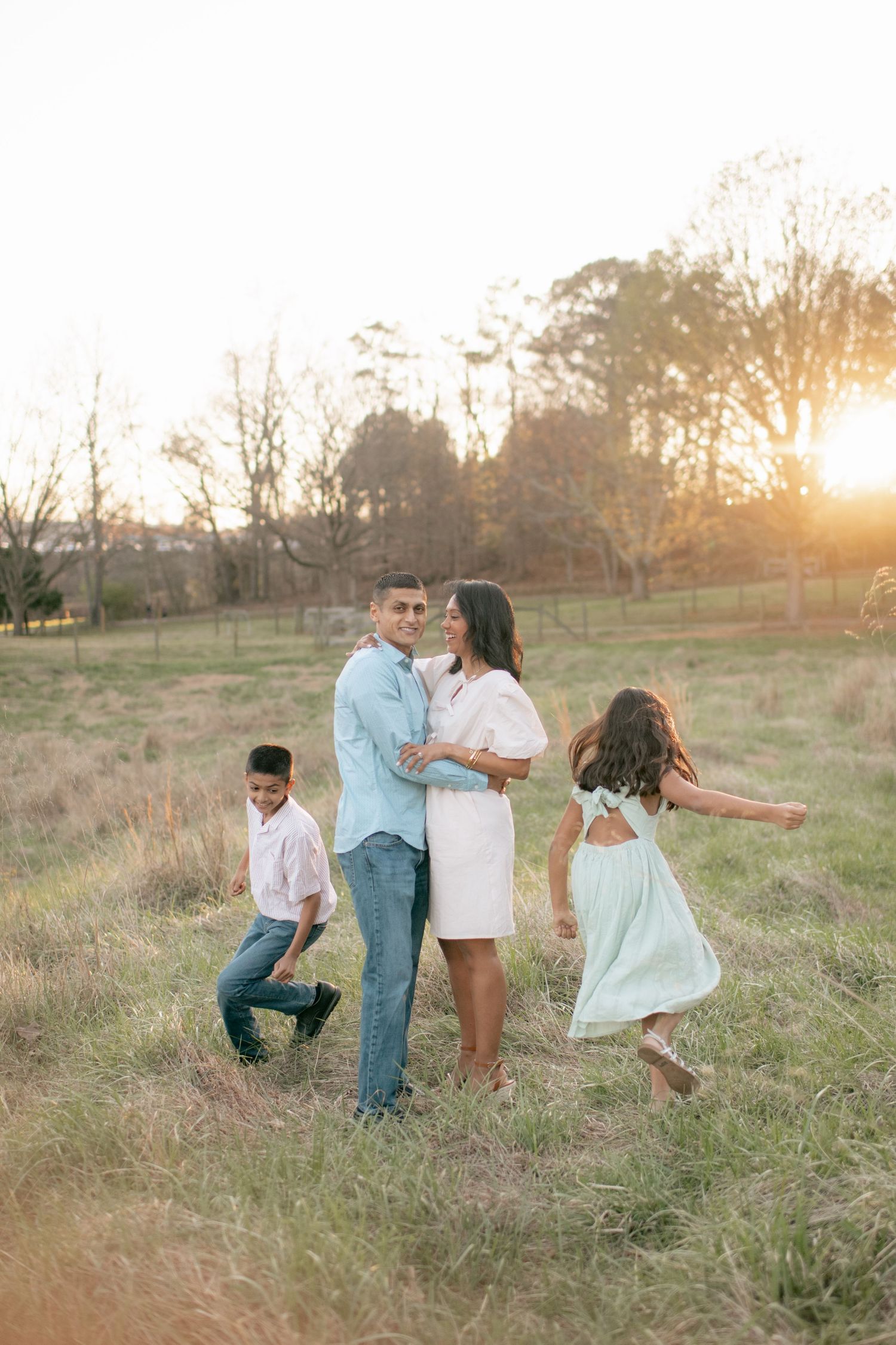 Family enjoying a playful moment together in a golden sunset field with tall grass and trees in the background.