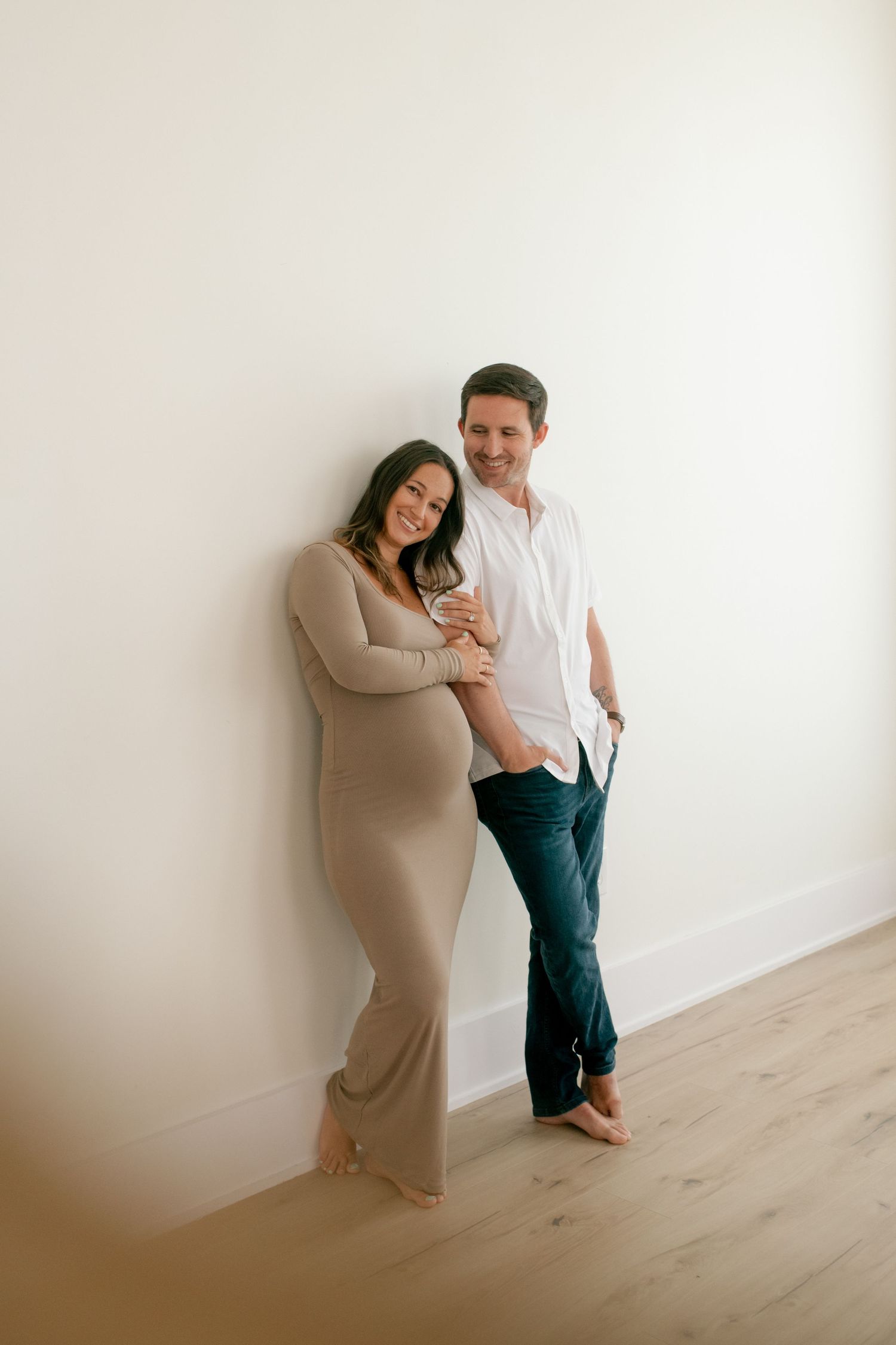 Expecting couple poses against white wall in casual attire for maternity photos.