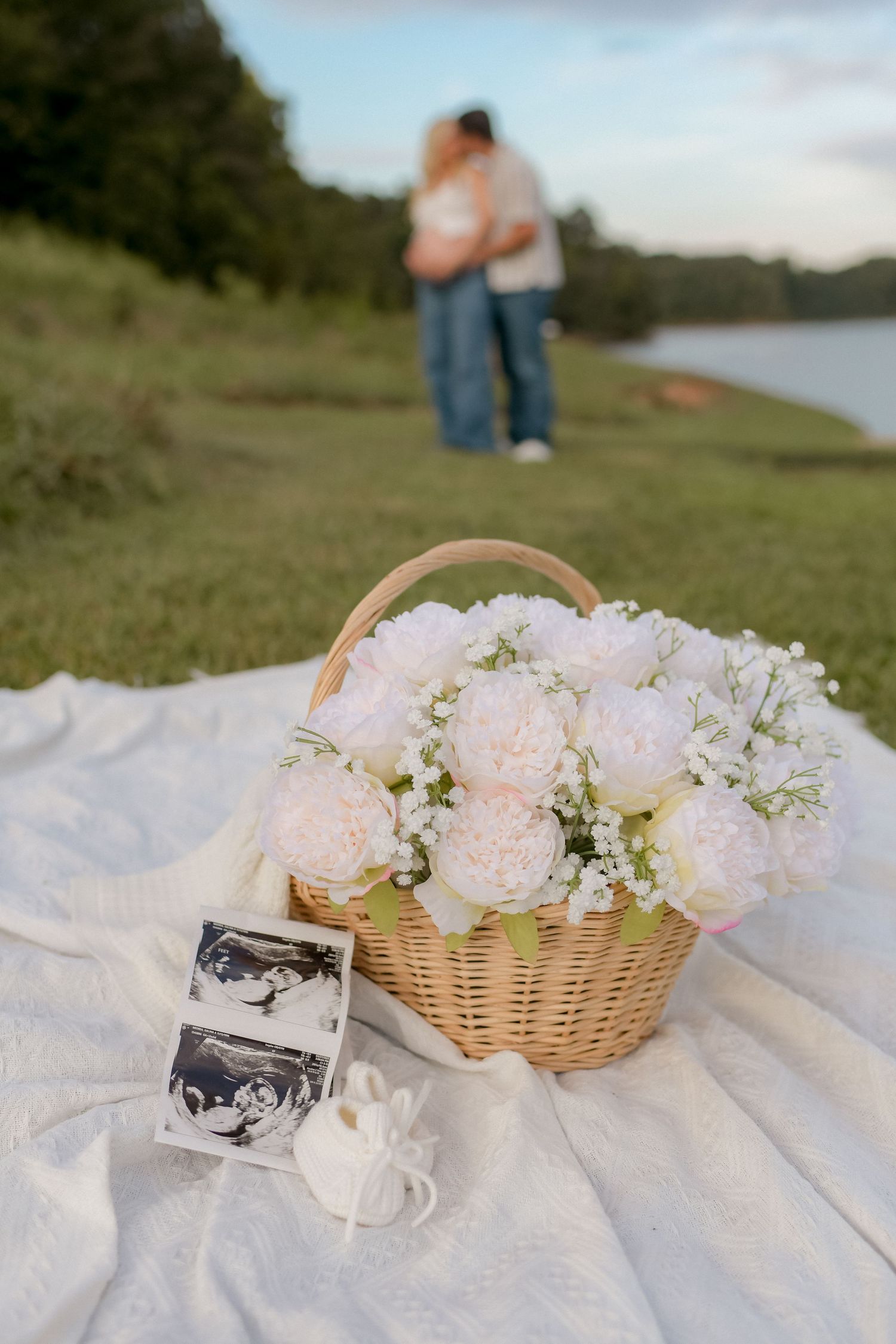 Wicker basket with pink and white flowers and ultrasound photo on white blanket outdoors.