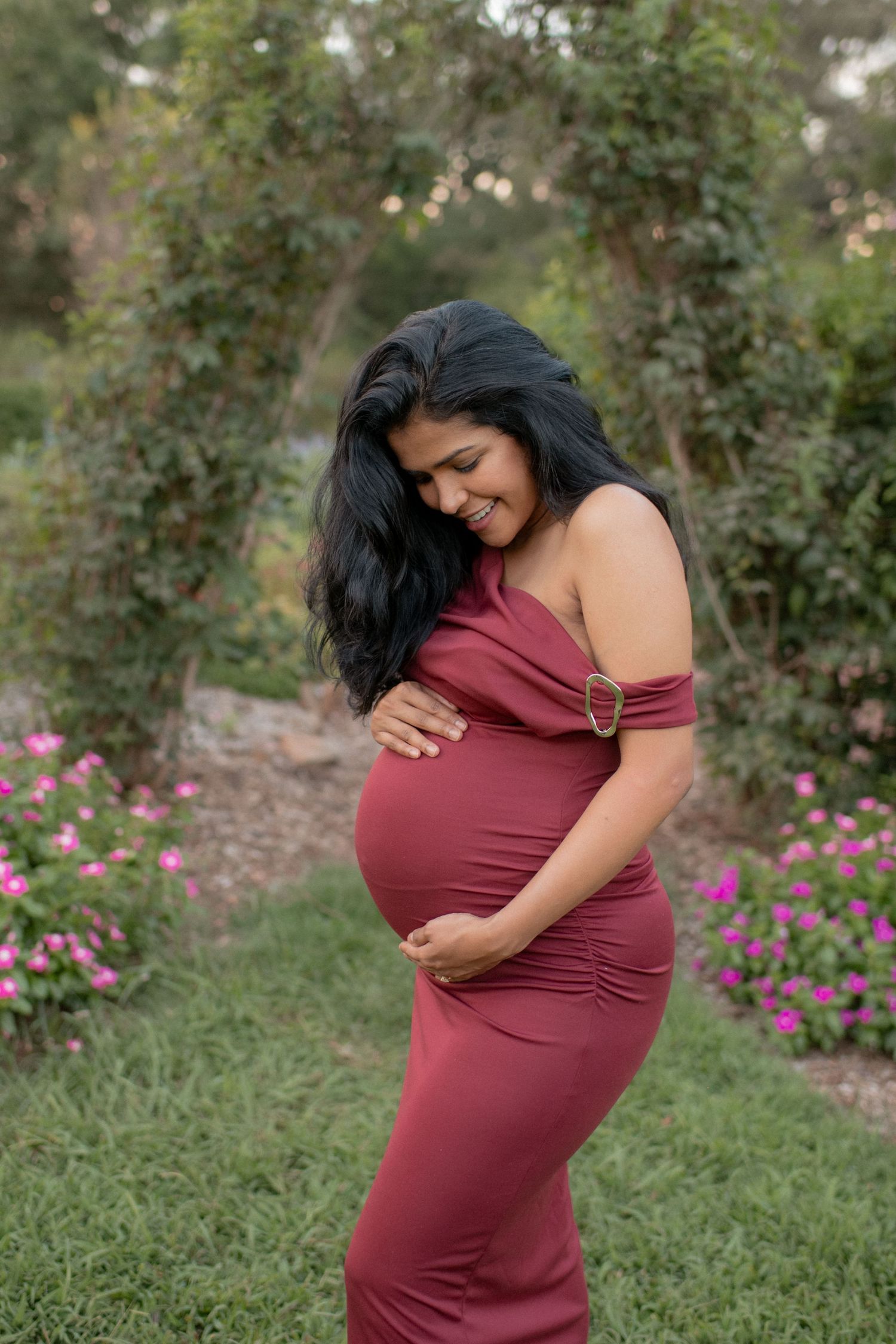 Maternity photos in a burgundy dress against a backdrop of flowering bushes.