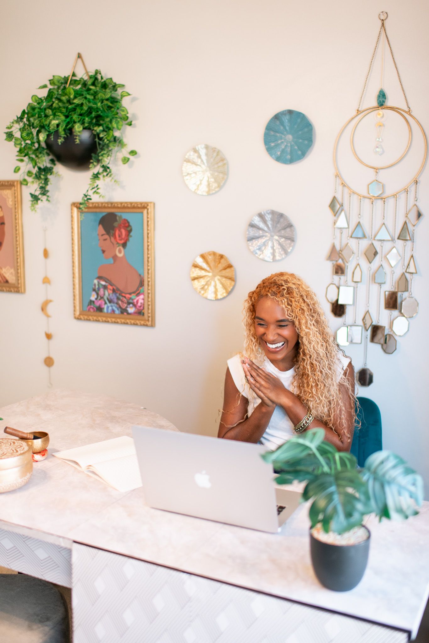 Smiling Black woman with curly blonde hair working on laptop at marble desk in boho home office with wall art and plants.
