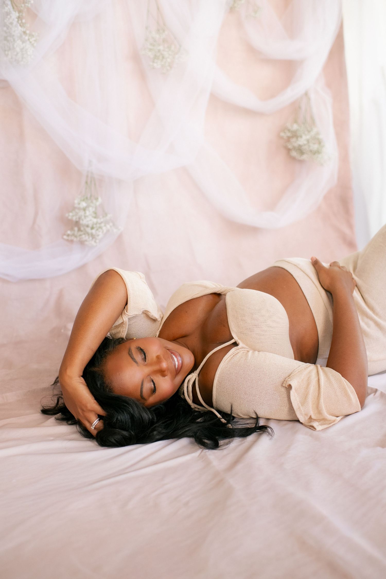 Relaxed maternity pose on pink fabric with cream-colored two-piece outfit.