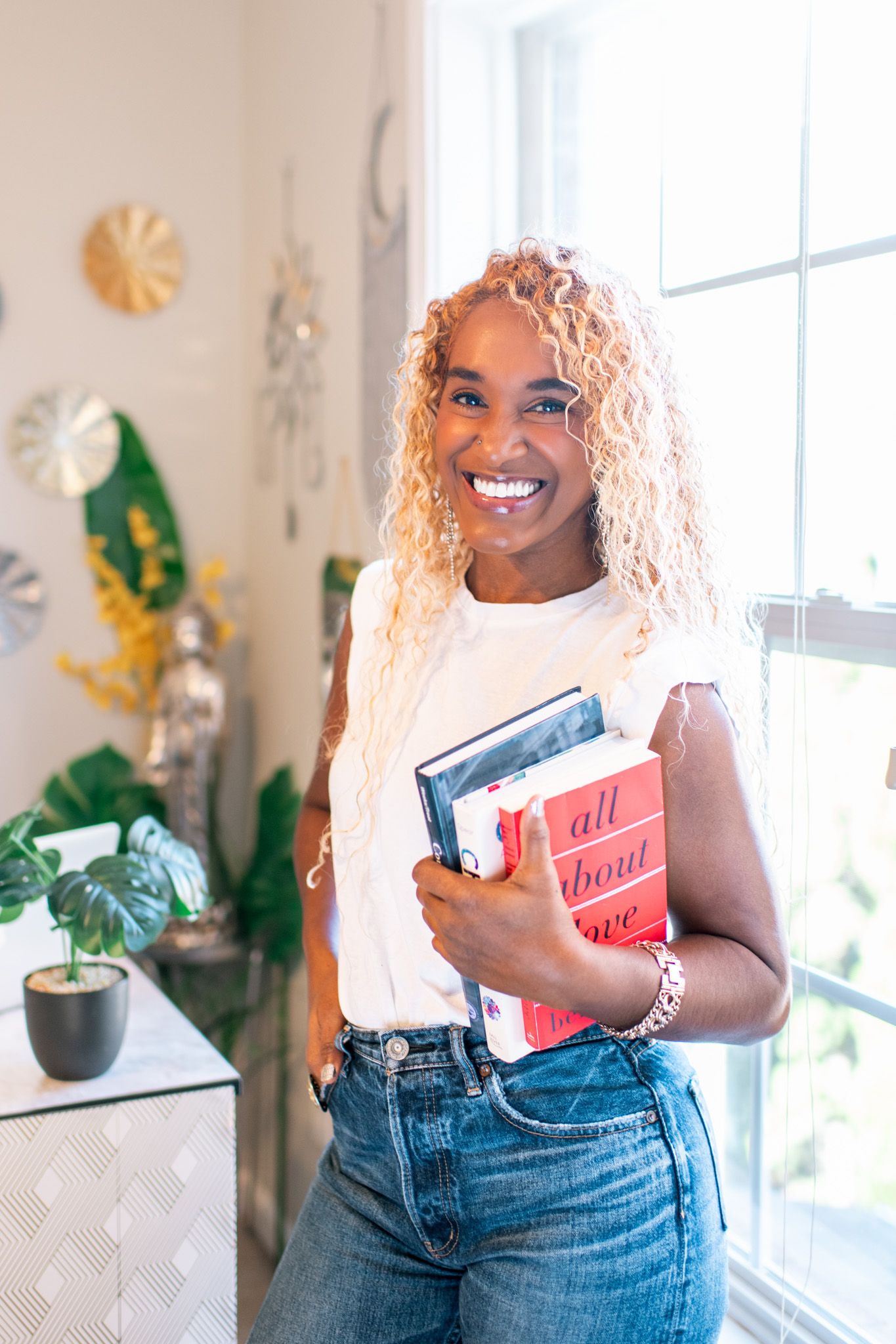 Person wearing denim jeans and white top holds book in bright sunlit room with decorative wall art.