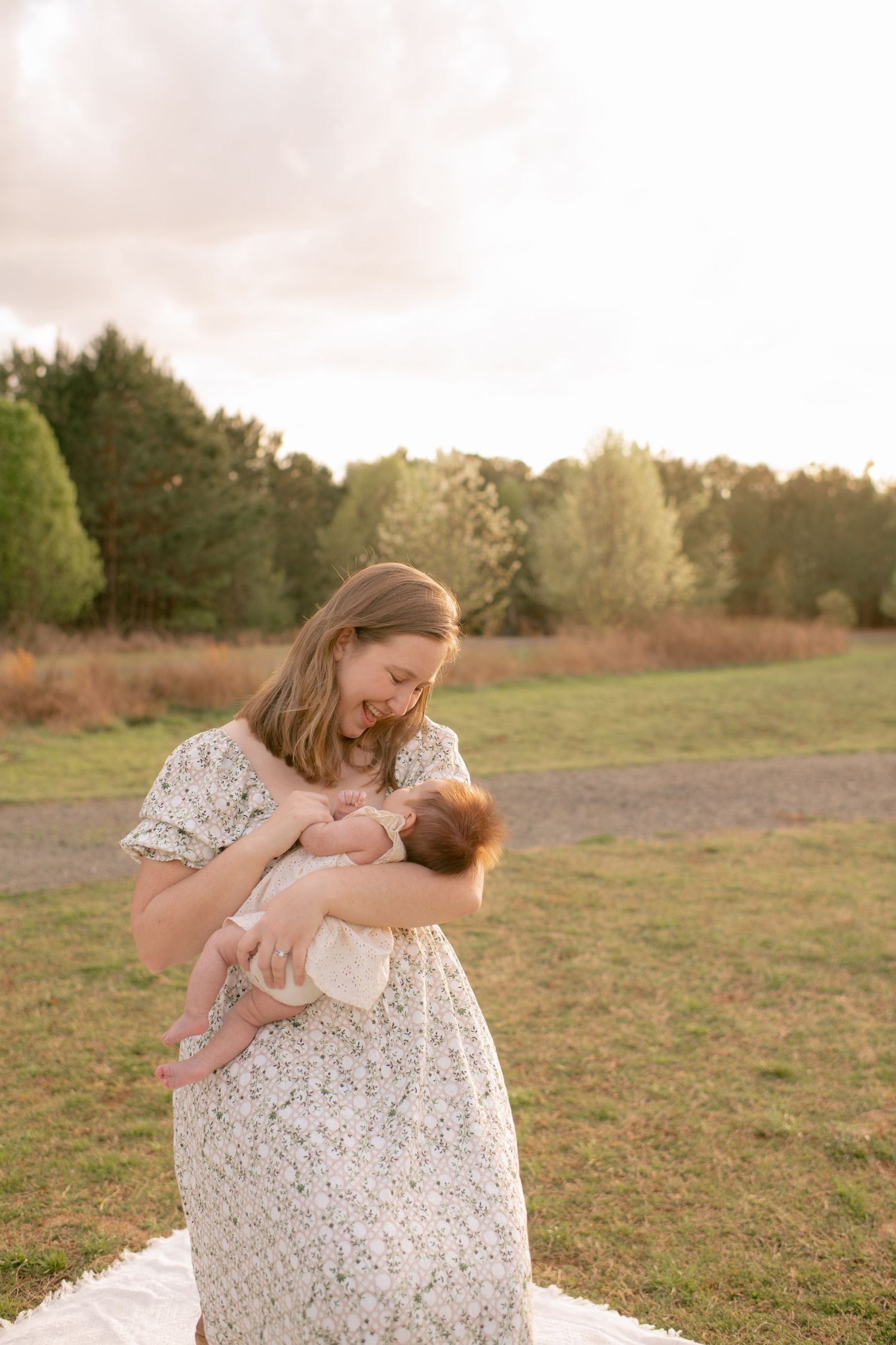 A tranquil nursing moment captured outdoors at golden hour with mother in a white lace dress on a grassy field.
