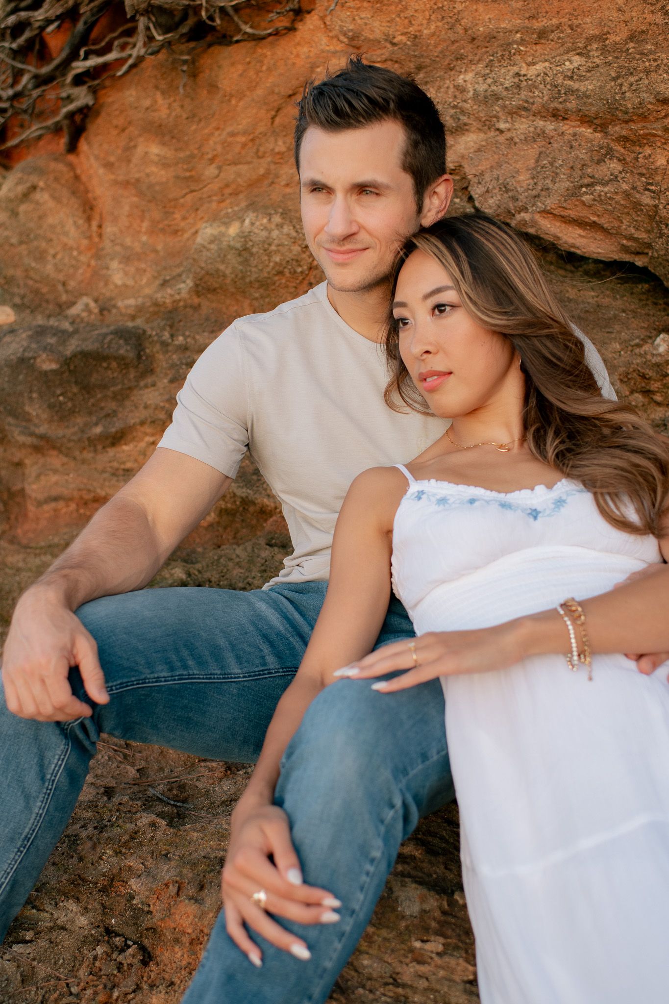 Romantic couple in casual attire sitting against red rock formation as wind blows through her hair.