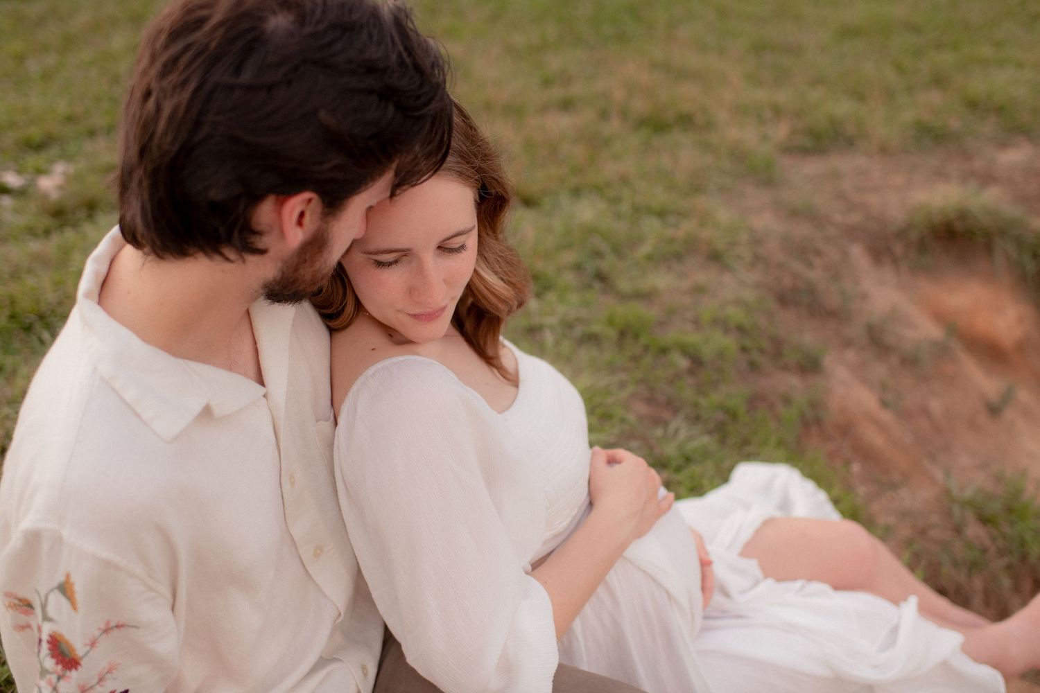 A romantic couple embraces in a grassy field during sunset while wearing white and beige outfits.