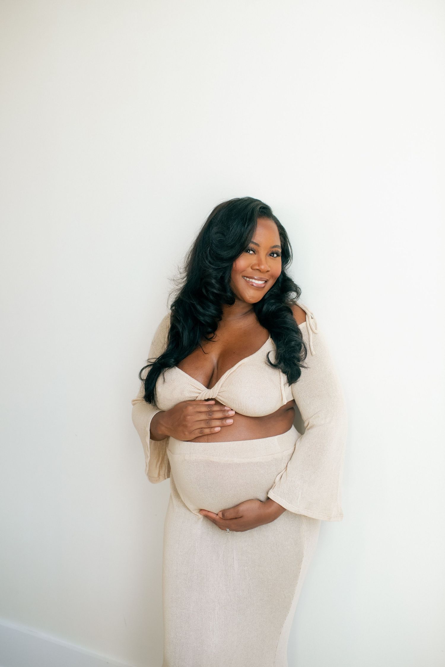 Maternity portrait against white wall showing expectant mother in cream bell-sleeve dress.