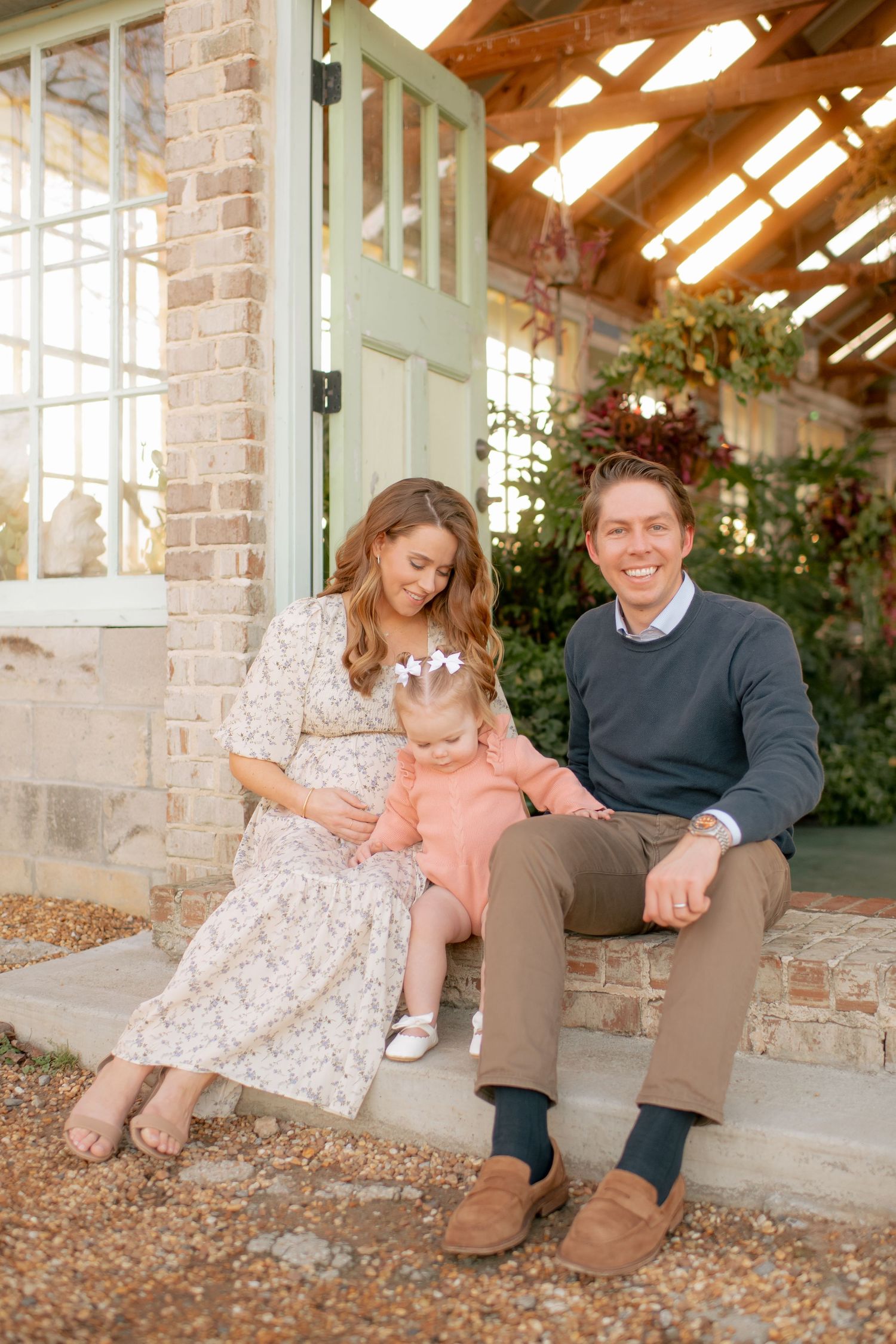 A family sits together on brick steps of a sunlit greenhouse with exposed wooden beams and climbing vines.