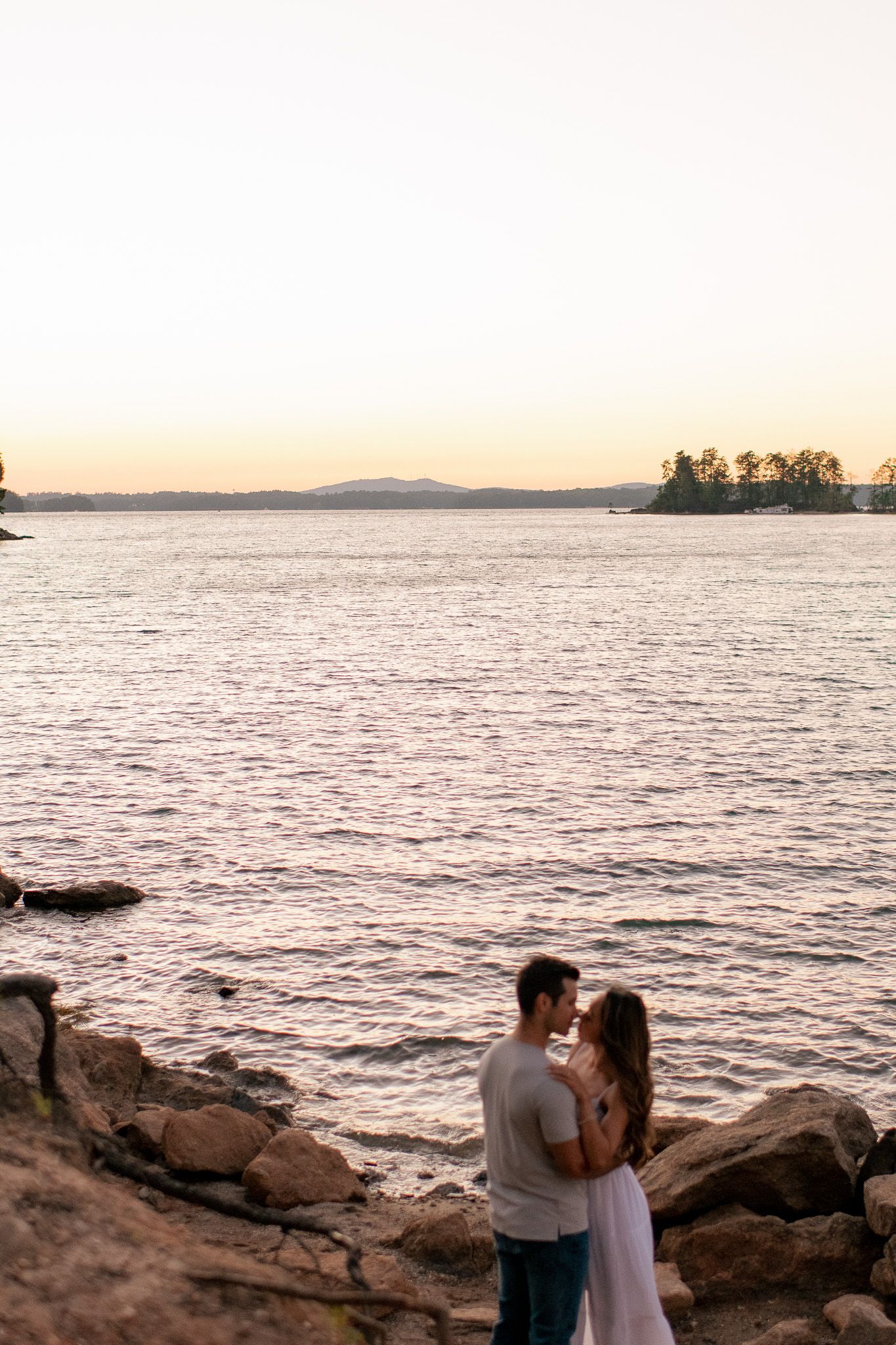 Scenic lakeside engagement photos on rocky shore during golden sunset with mountains in the distant background.