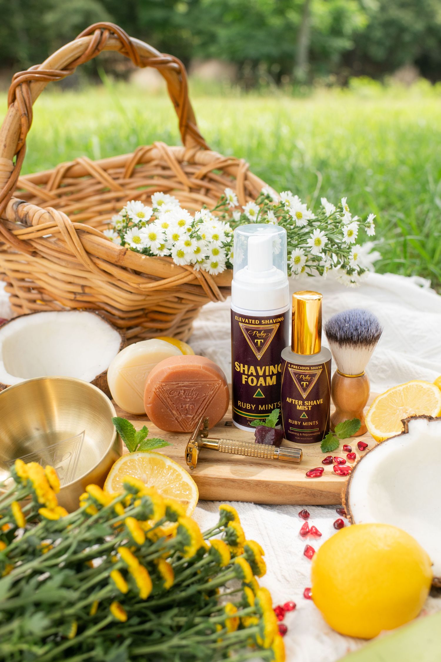 Shaving foam and after-shave mist displayed outdoors with fruits, flowers, and a wicker basket on a picnic blanket.
