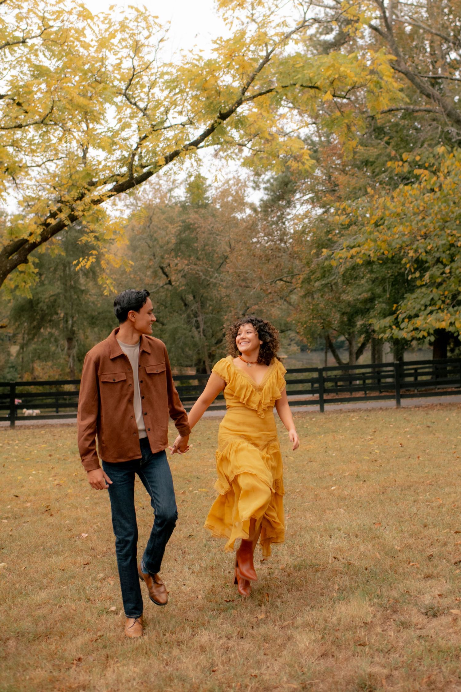 Playful couple running through autumn field with yellow leaves and wooden fence in background.