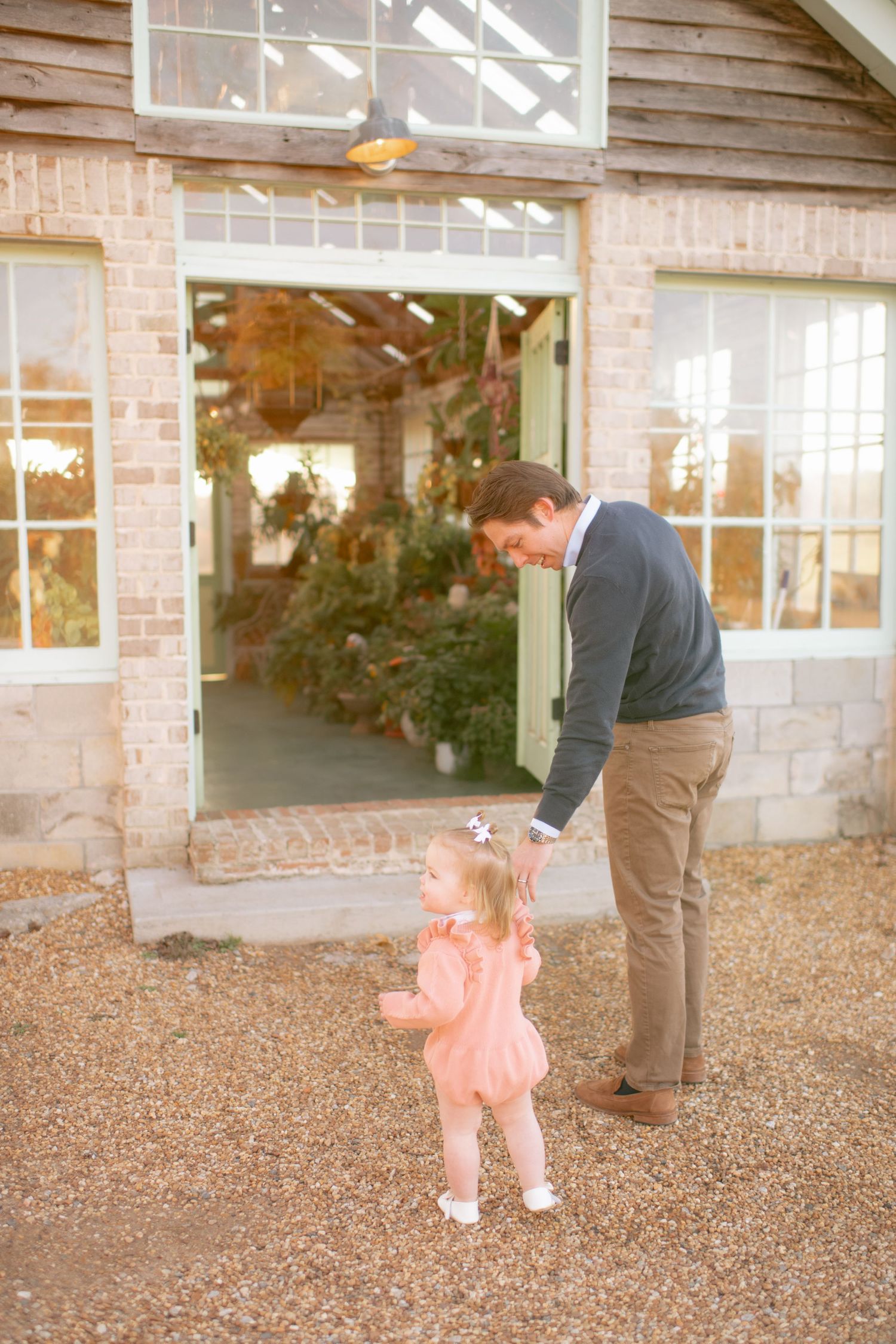 A tender outdoor moment between a child in a pink outfit and an adult near a rustic building entrance.