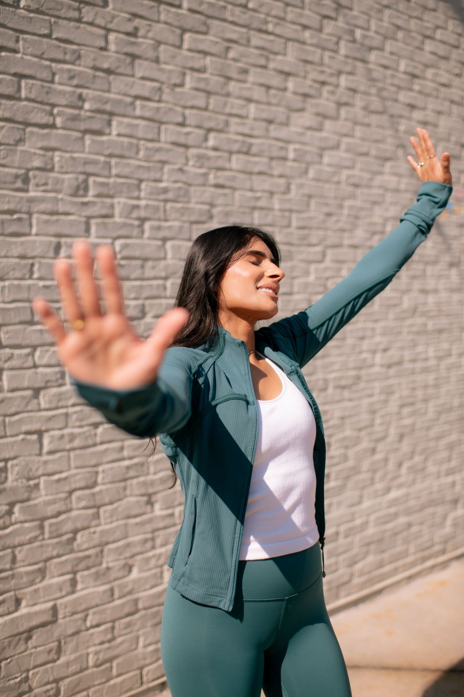 Woman in teal athletic wear stretches joyfully with arms raised against brick wall.
