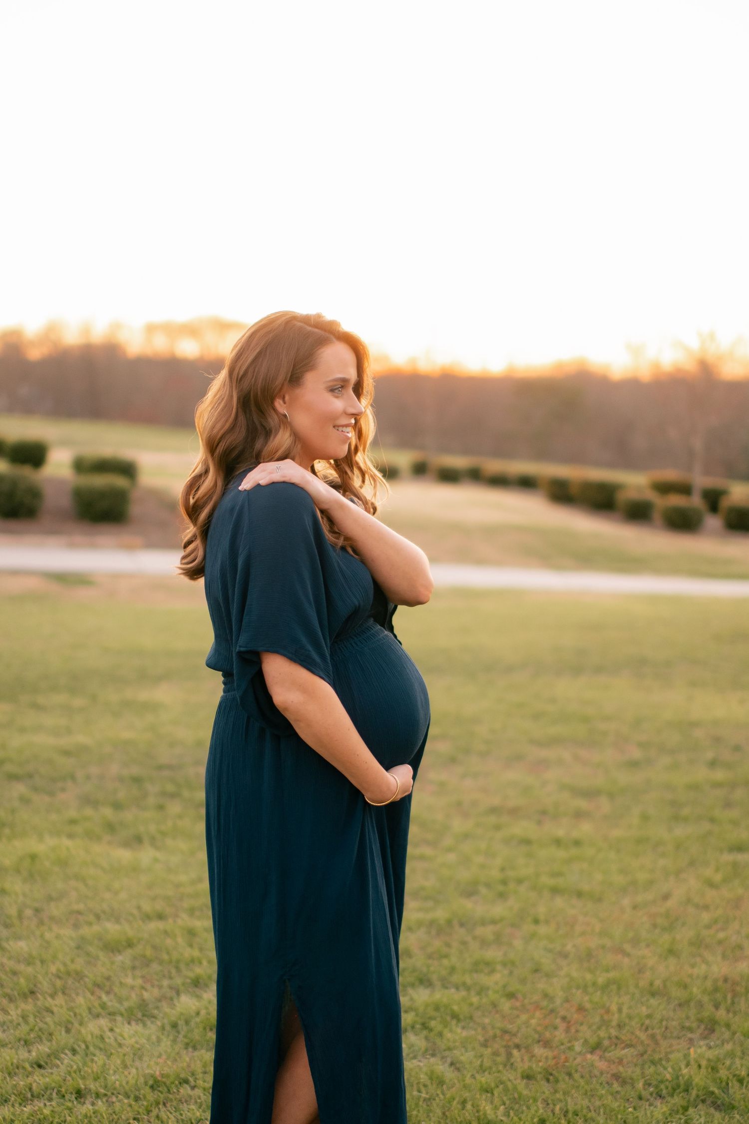 A pregnant person in a dark blue dress stands in a grassy field at sunset while tenderly cradling their belly.