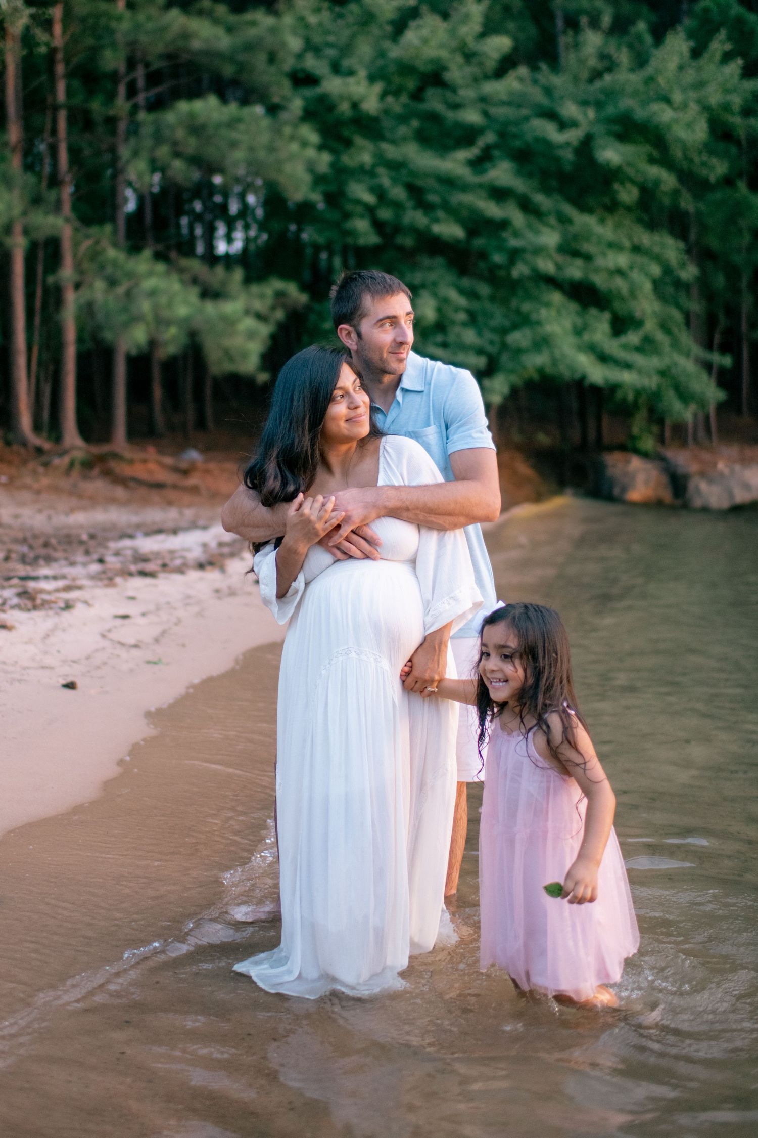 Family wearing white and pink dresses poses for tender moments by the water's edge at a scenic beach.