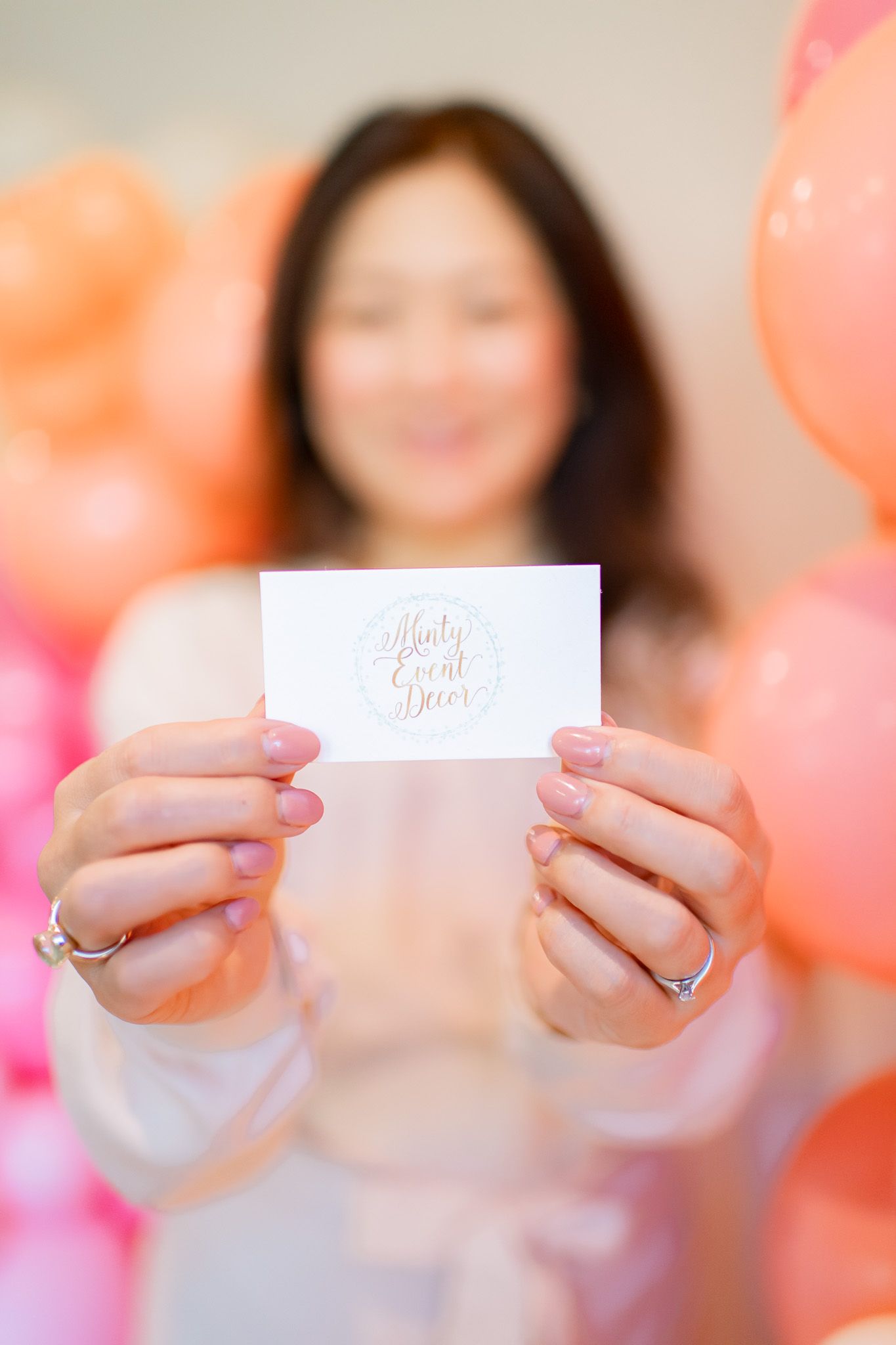 Hands holding white name cards against a blurred coral and pink balloon background.