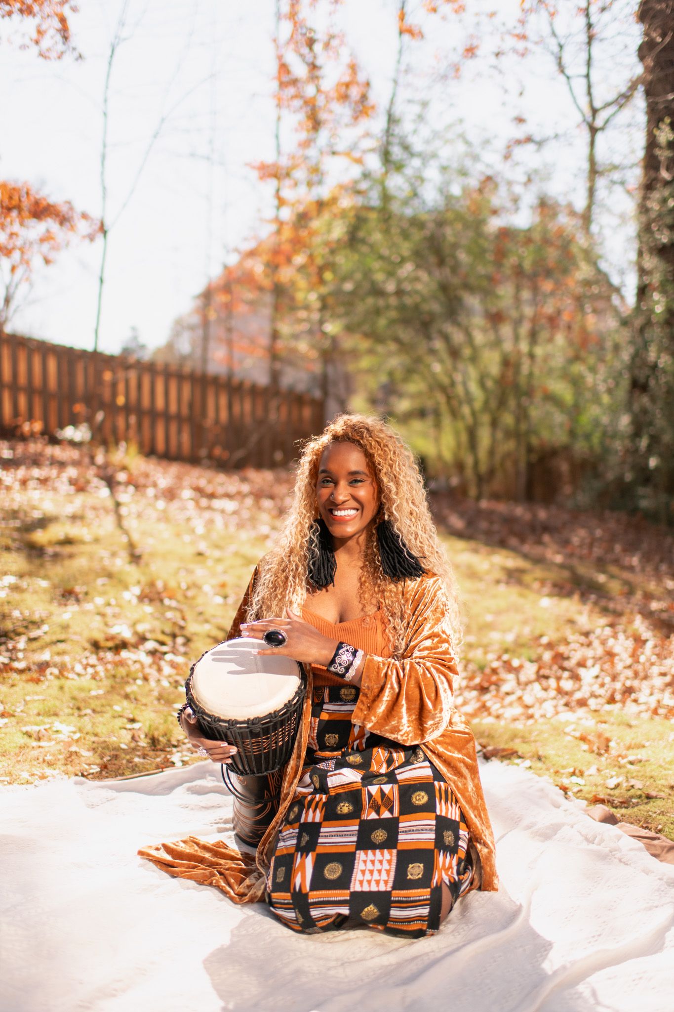 Smiling woman with curly hair playing a djembe drum outdoors in autumn, wearing a colorful African print skirt.
