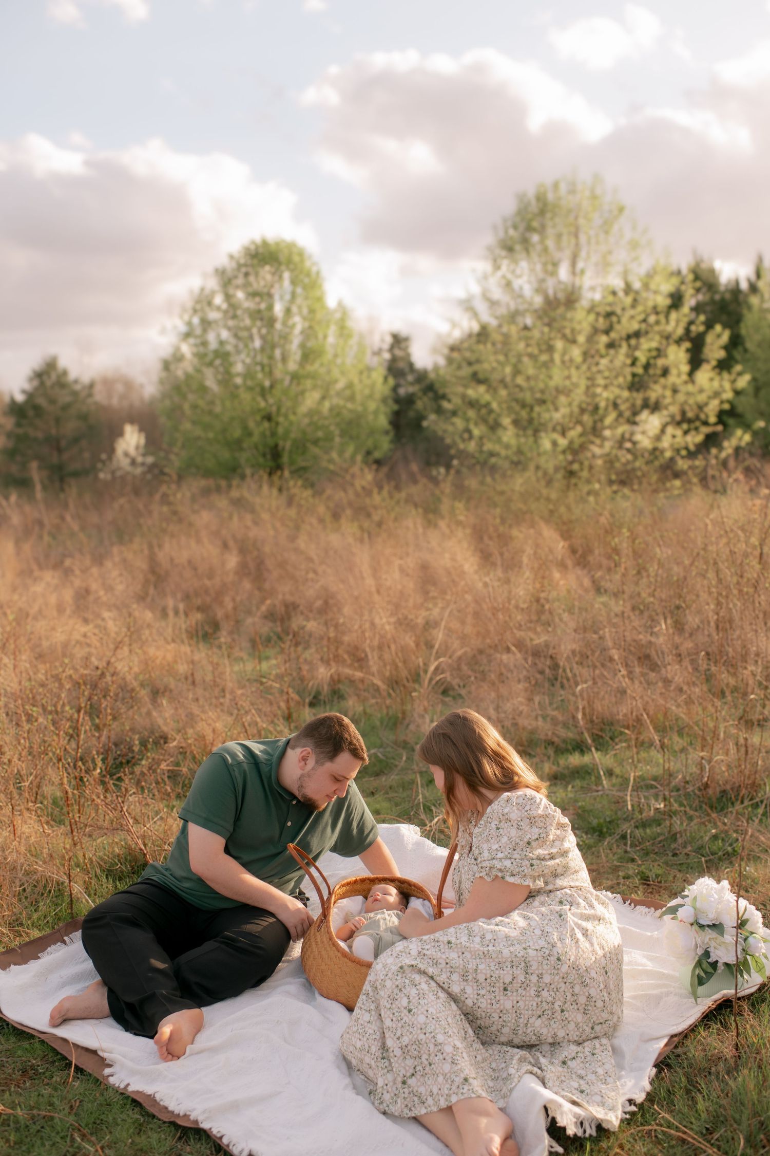Couple enjoying a romantic spring picnic on white blanket in field with tall golden grass and green trees.
