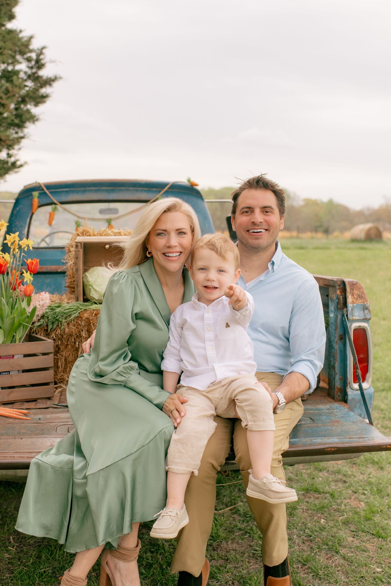 Family sits together on vintage turquoise pickup truck bed with hay bales and wooden crates in rustic outdoor setting.