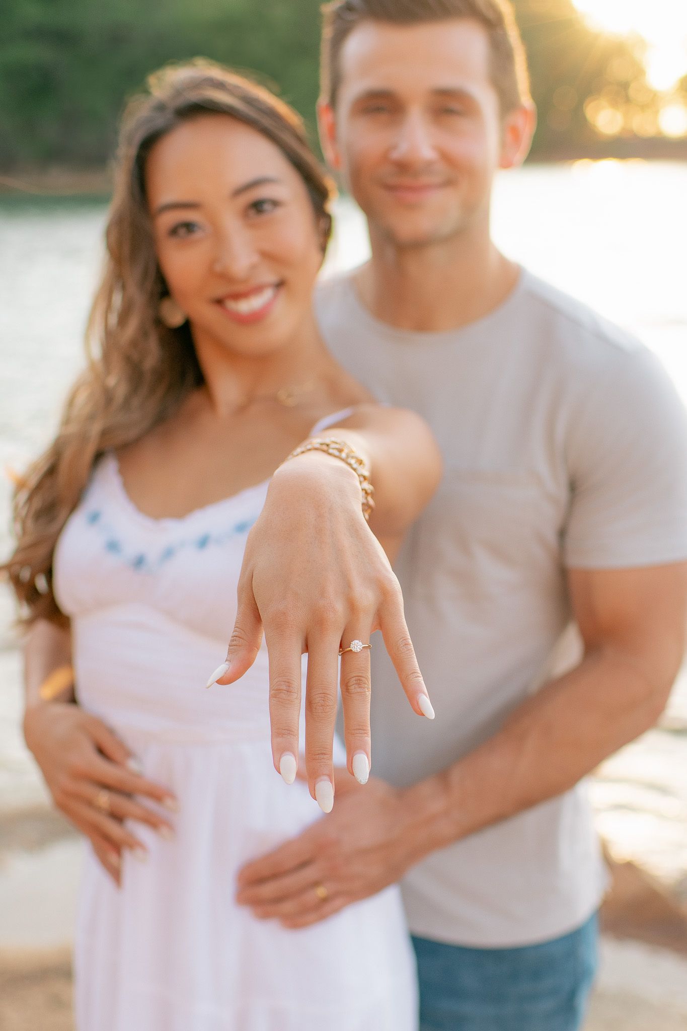 Close-up of engaged couple at sunset with woman showing engagement ring on her hand while wearing a white dress.