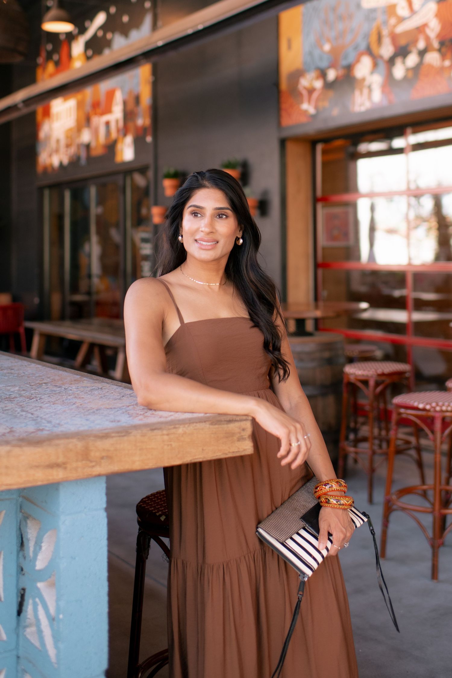 A woman in a brown dress poses at a rustic restaurant bar with wooden stools and warm lighting.