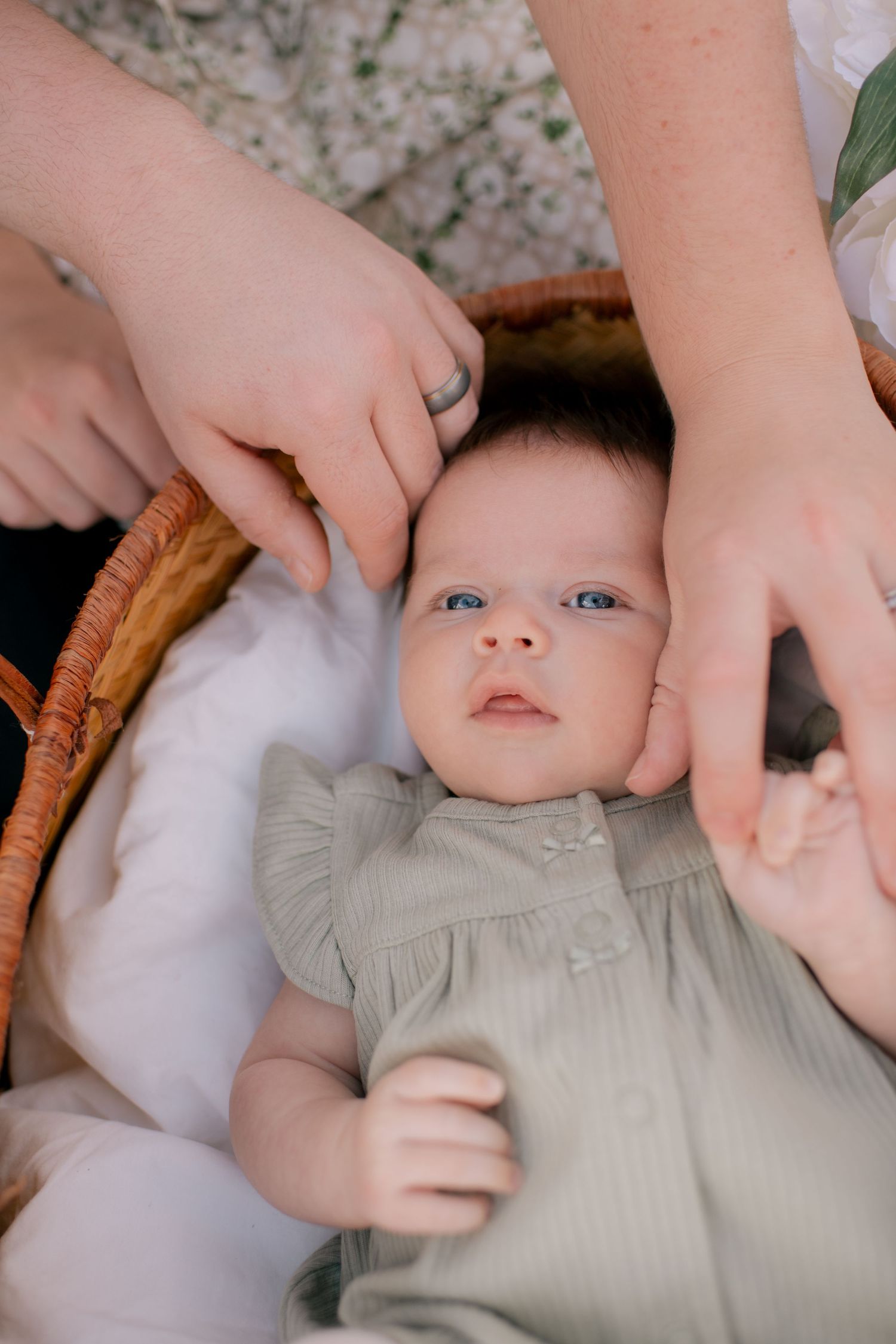 A small baby wearing a light gray outfit lies in a wicker basket while hands gently cradle their head.