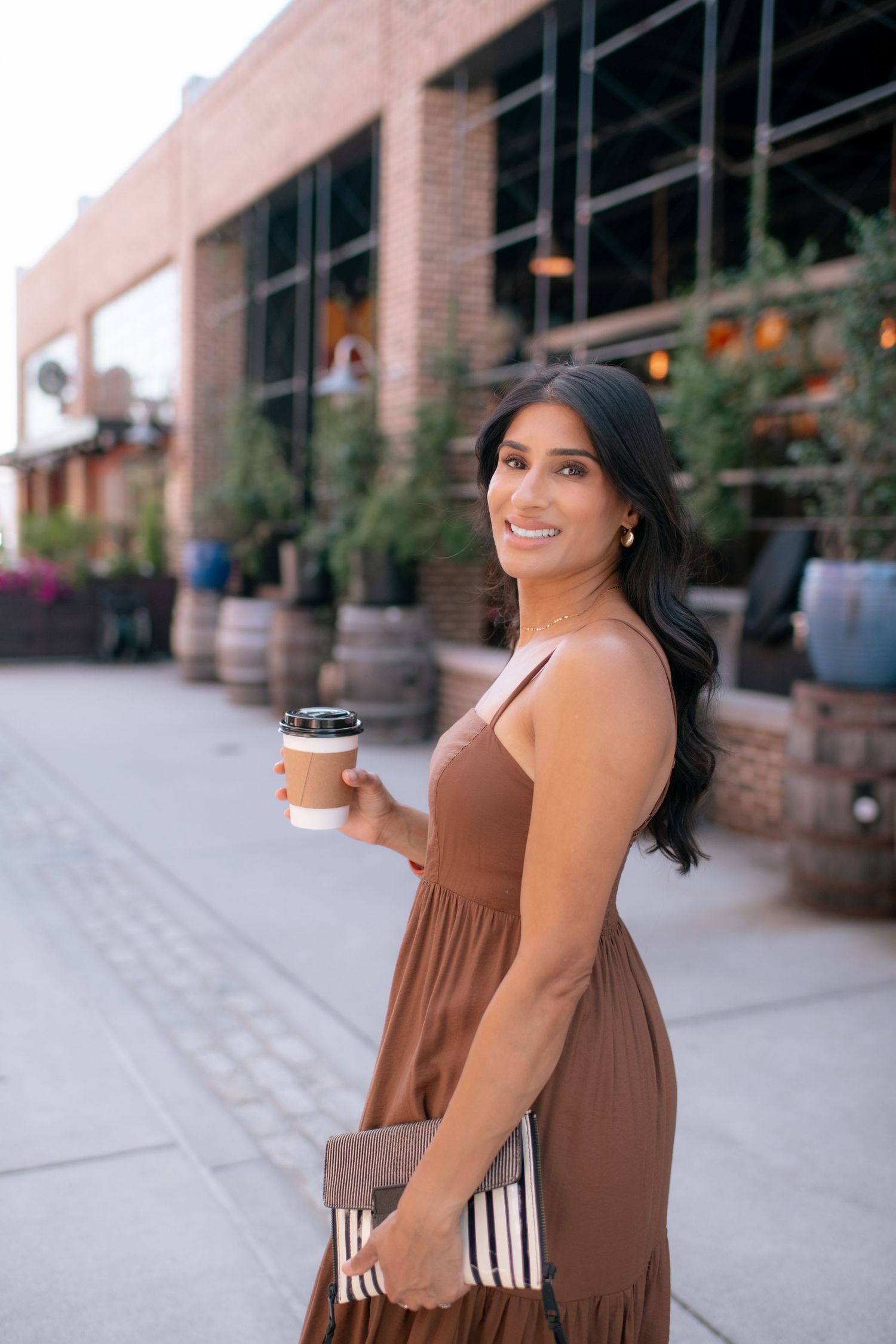 Person in brown dress enjoying coffee while sitting outdoors near brick buildings on a sunny day.