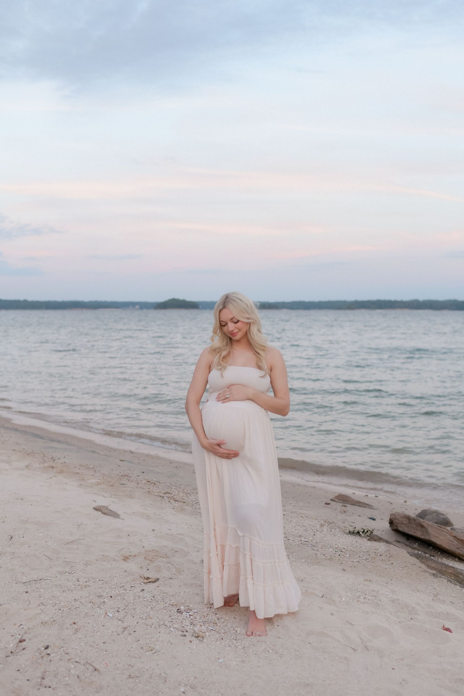 A woman in a flowing dress cradles her baby bump on a serene beach at sunset.