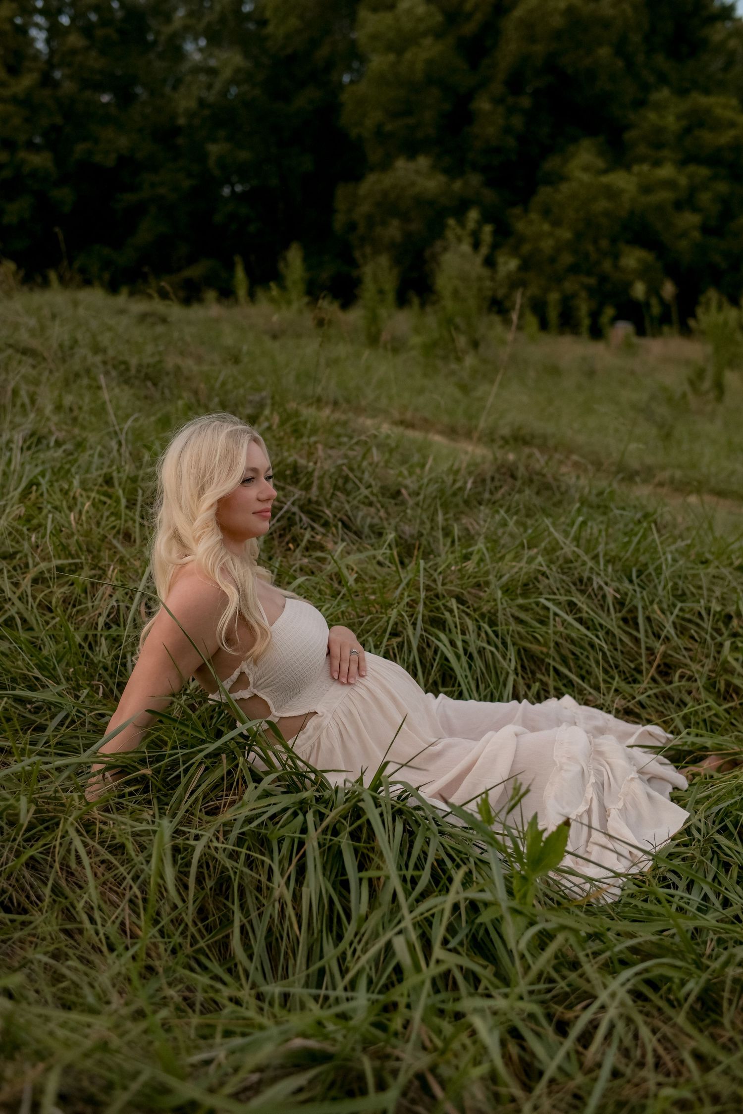 An expectant mother poses elegantly in a light dress among tall grass during golden hour.