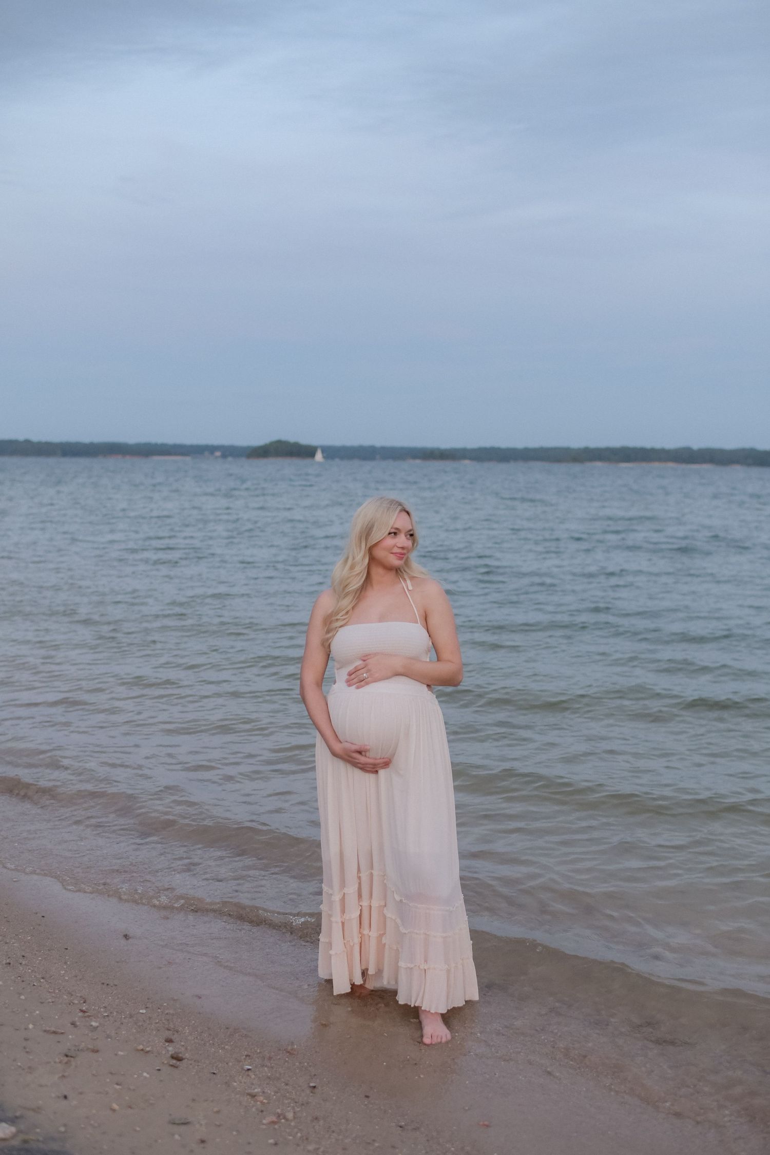 A woman in a light dress stands on a beach near the ocean.