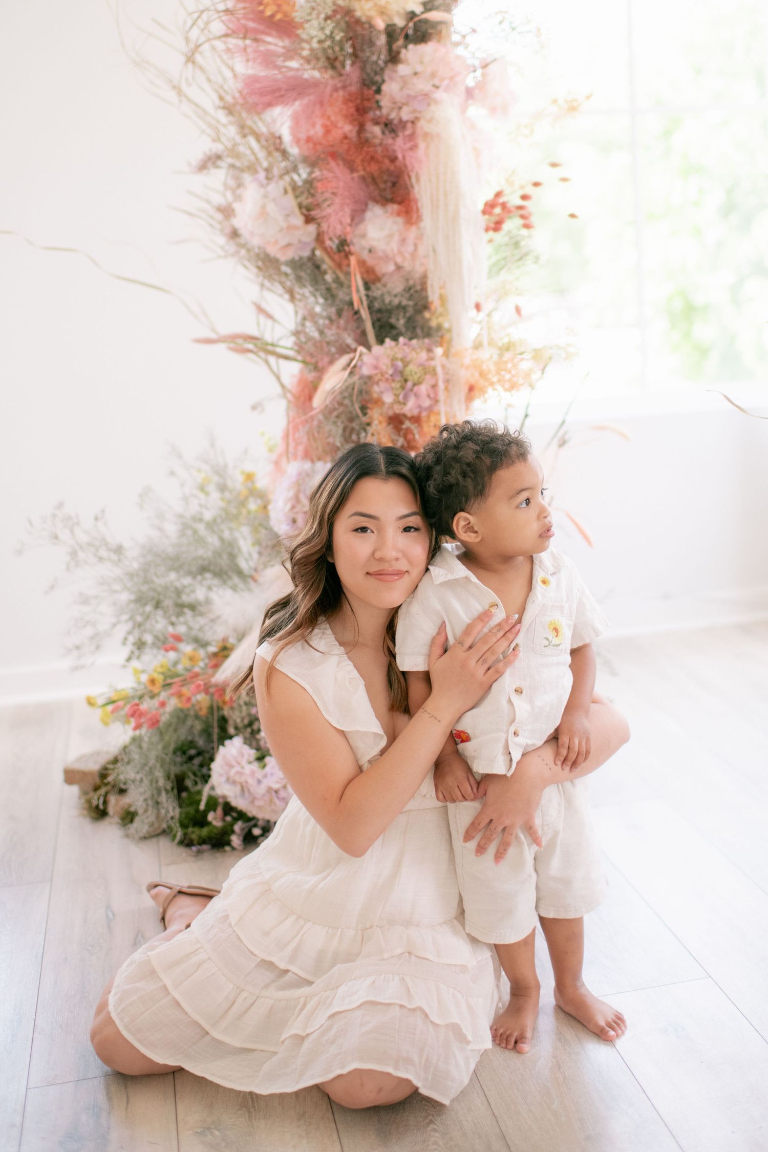 A tender portrait of parent and child in matching white outfits against a backdrop of pink and white floral arrangements.