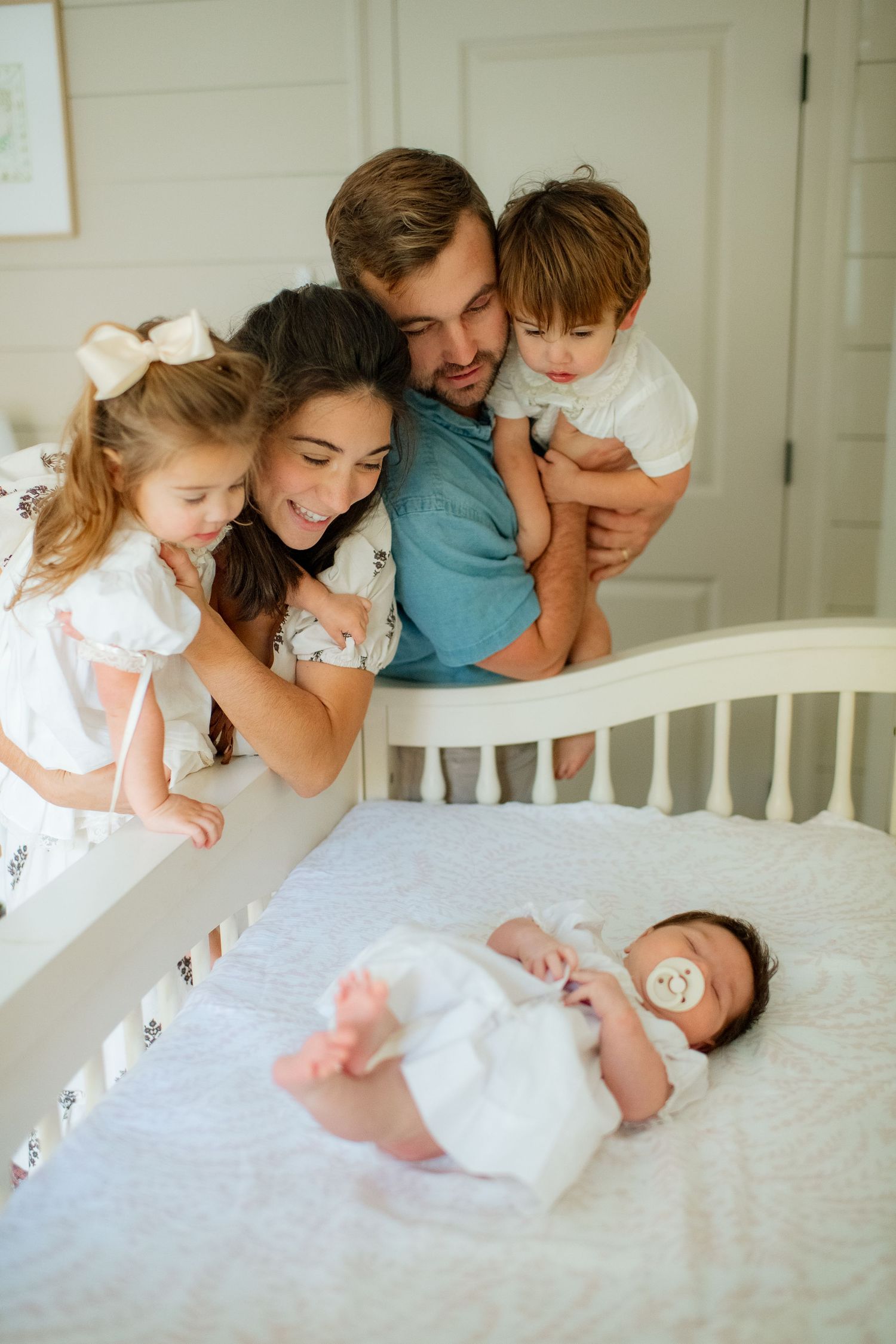 Family leaning over a white crib to admire their newborn baby together.