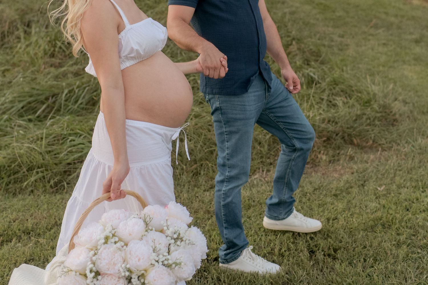 Couple walking through tall grass during outdoor maternity session with flowers.