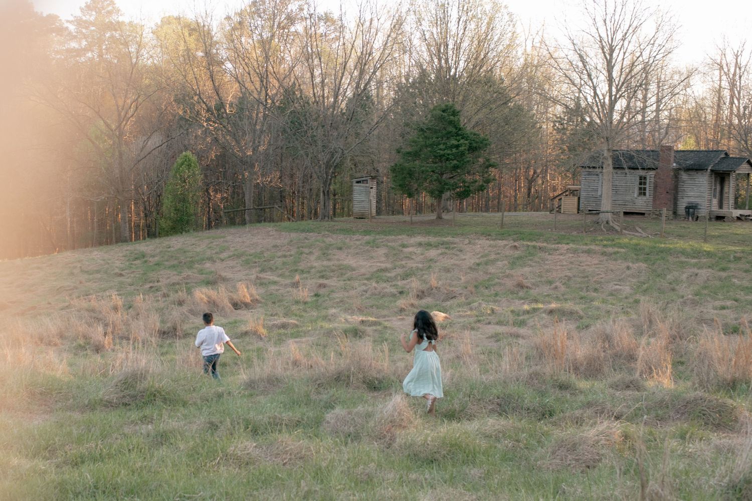 Two children playing in an overgrown field at sunset with trees and old buildings in the background.