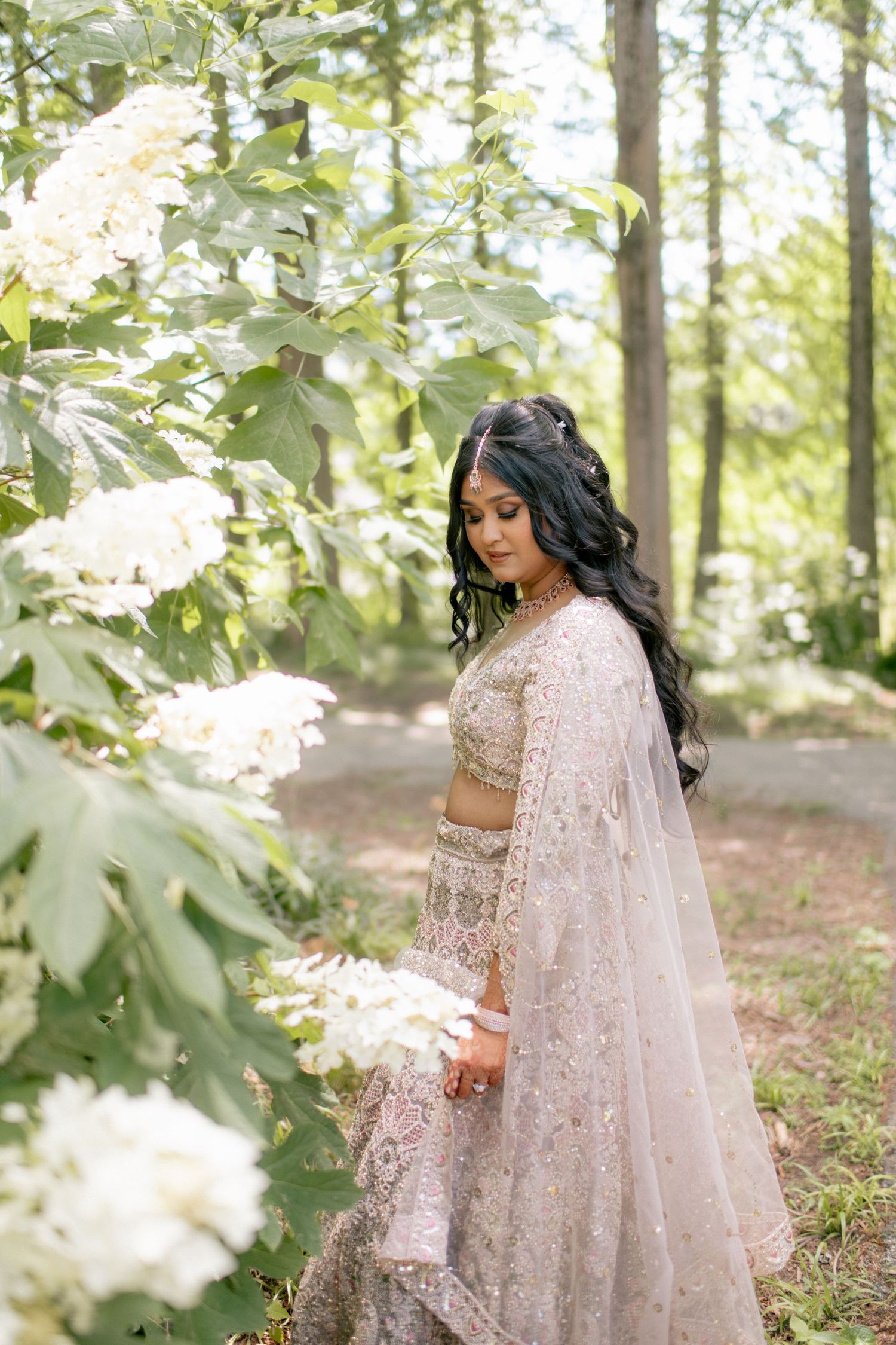 Traditional South Asian bridal lehenga in blush pink with delicate embroidery photographed in a dreamy outdoor forest setting.