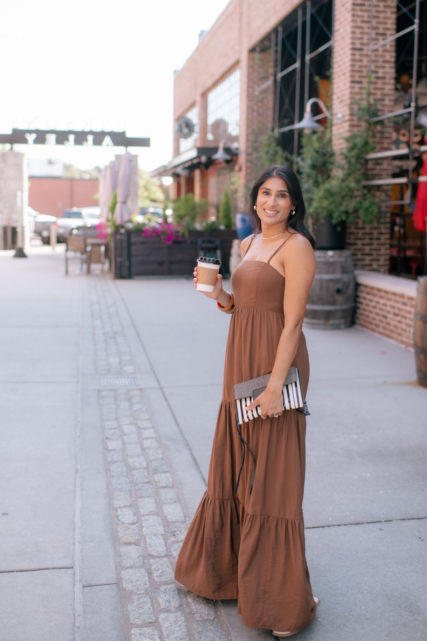 Woman in brown maxi dress holding coffee and striped clutch on brick-lined street.