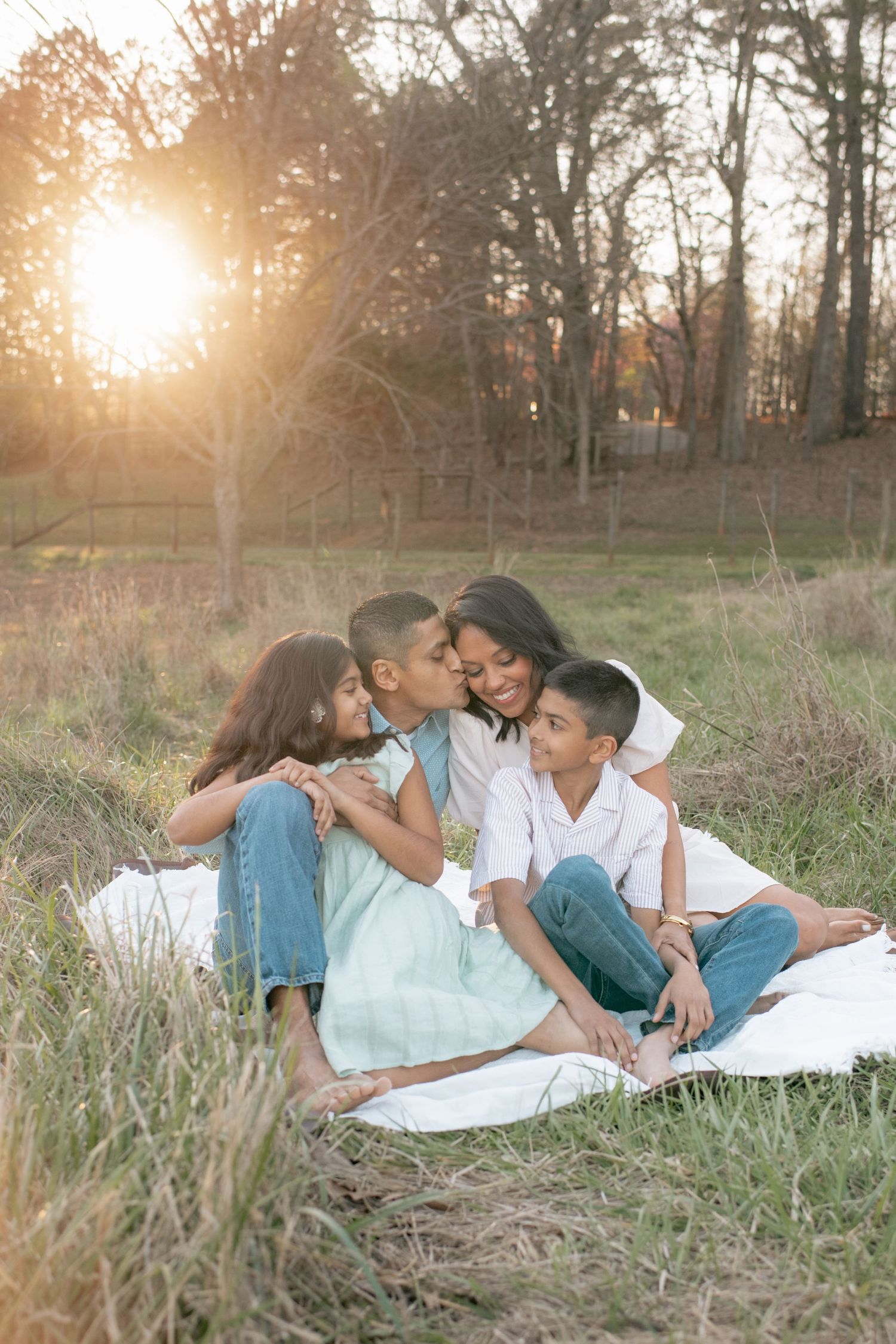 A tender family moment captured at sunset on a white blanket in a grassy field surrounded by trees.