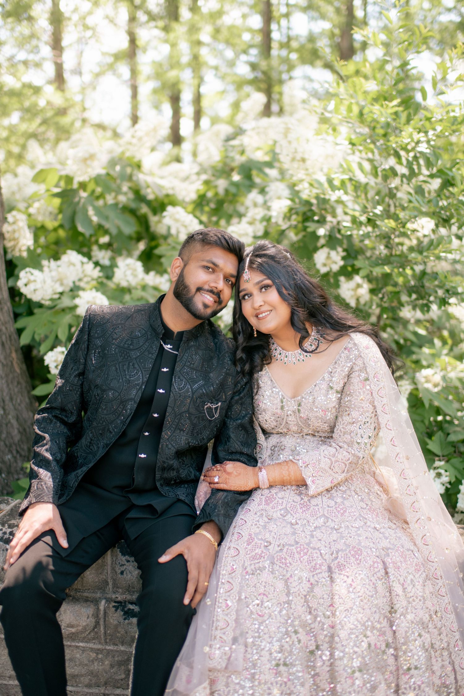 A couple sits together on stone steps surrounded by white flowering bushes in a forest setting during a photo session.