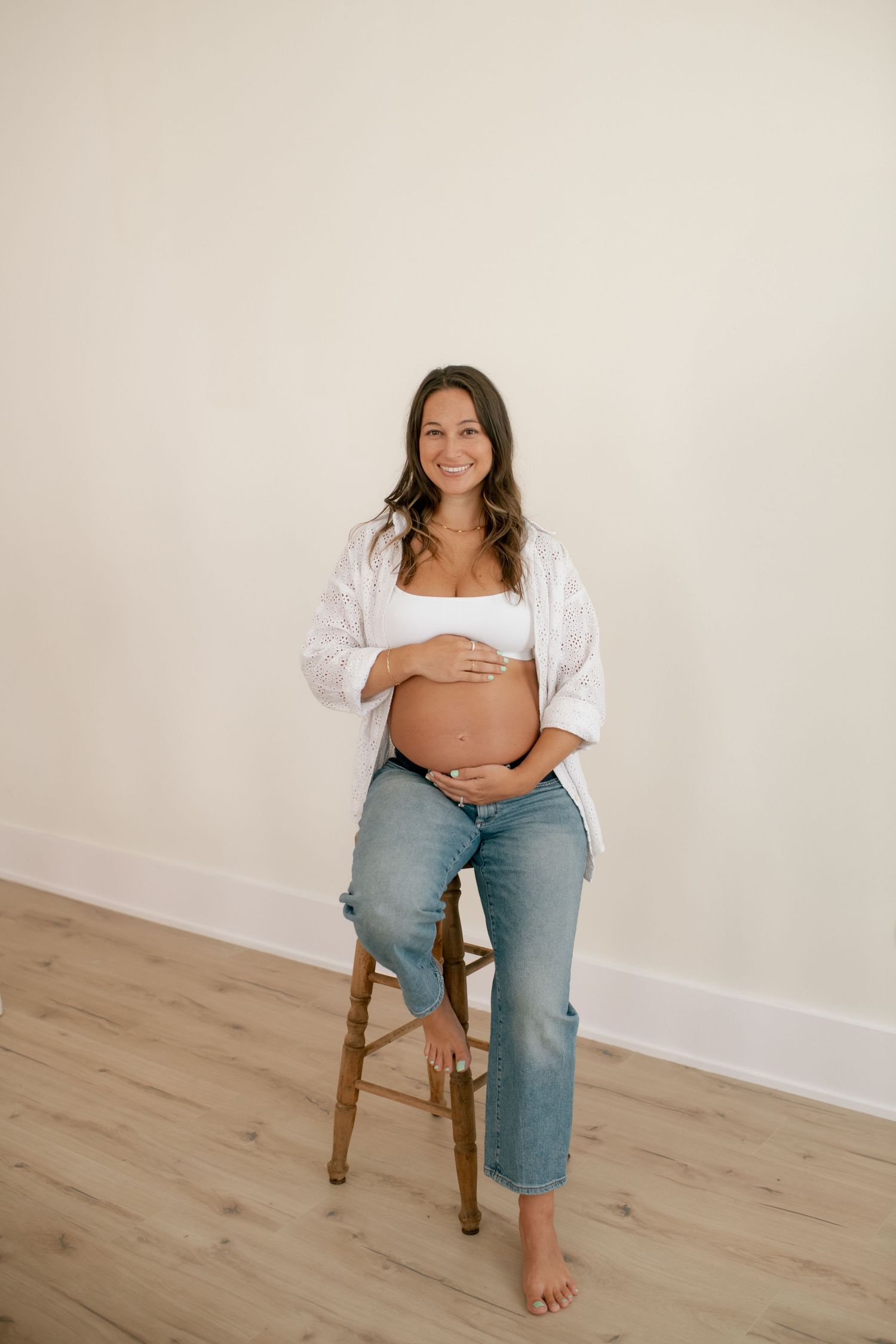 Casual maternity portraits of expectant mother sitting on wooden stool by white wall.
