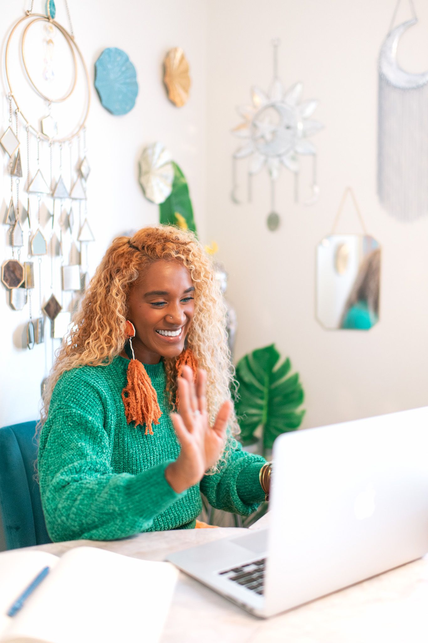 Smiling Black woman with curly blonde hair waving at laptop in boho home office with dream catchers on wall.