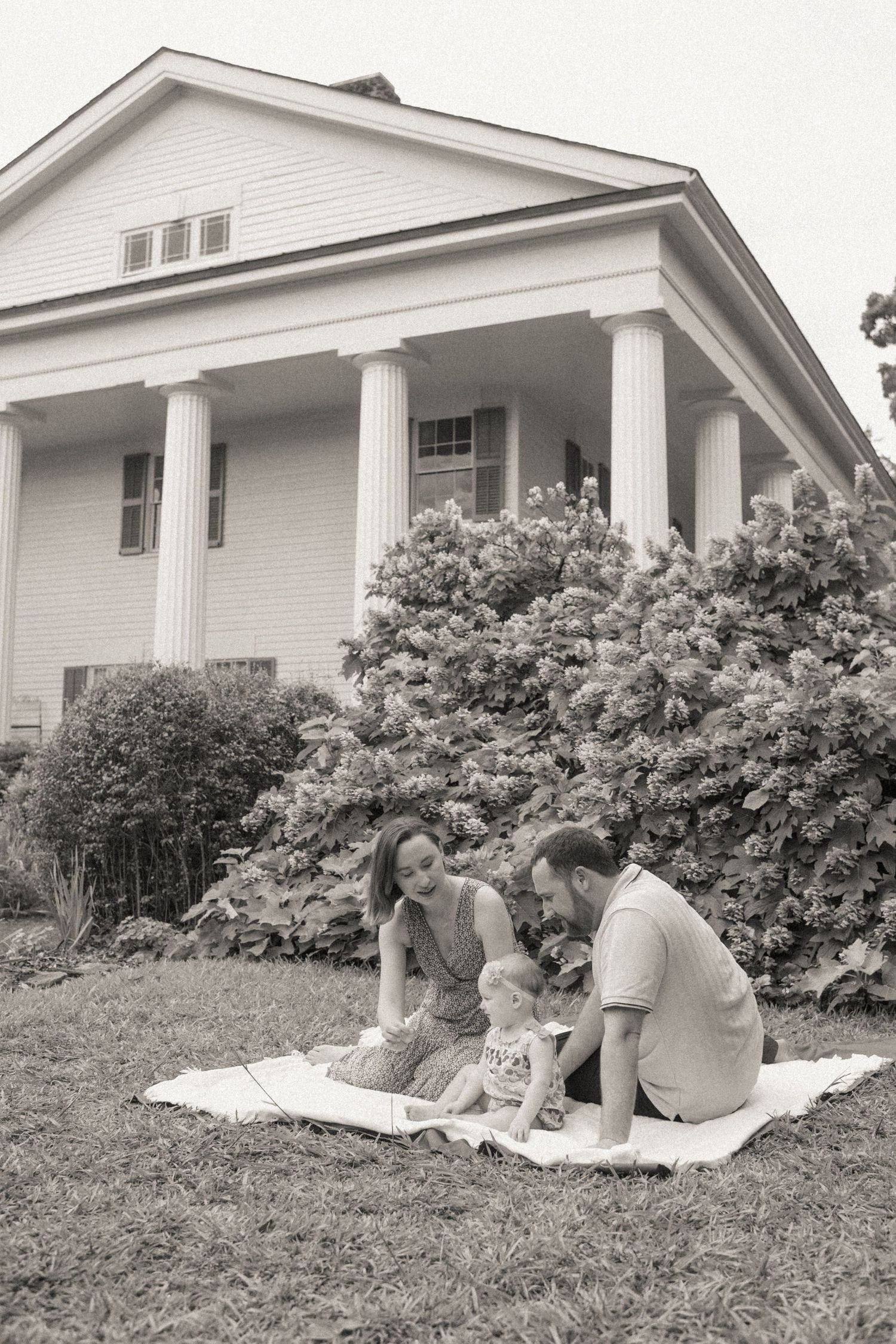 A couple sits on a blanket for a picnic in front of a white colonial-style house with tall columns.