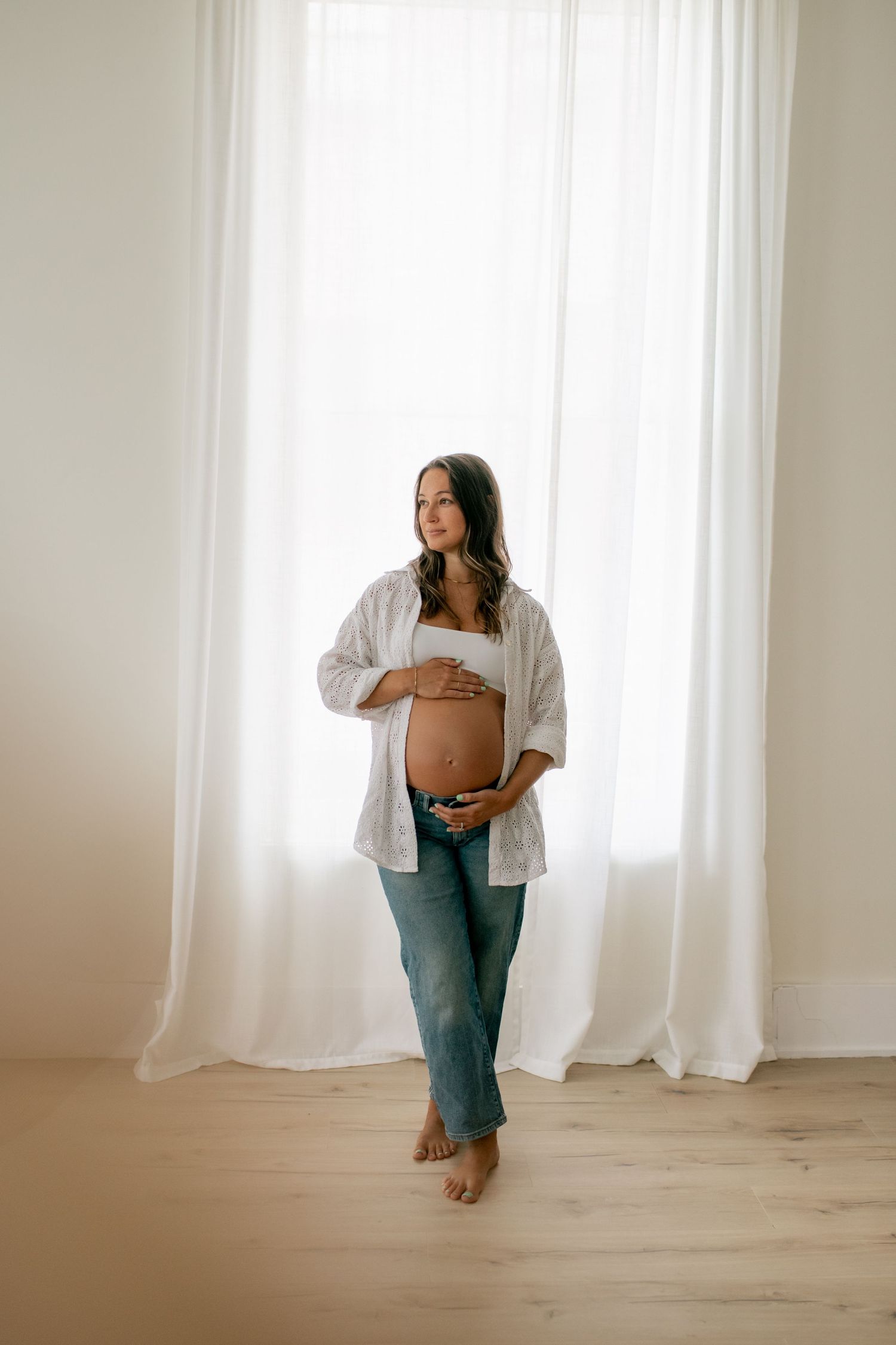 Expectant mother in jeans and white top poses by window in minimalist setting.