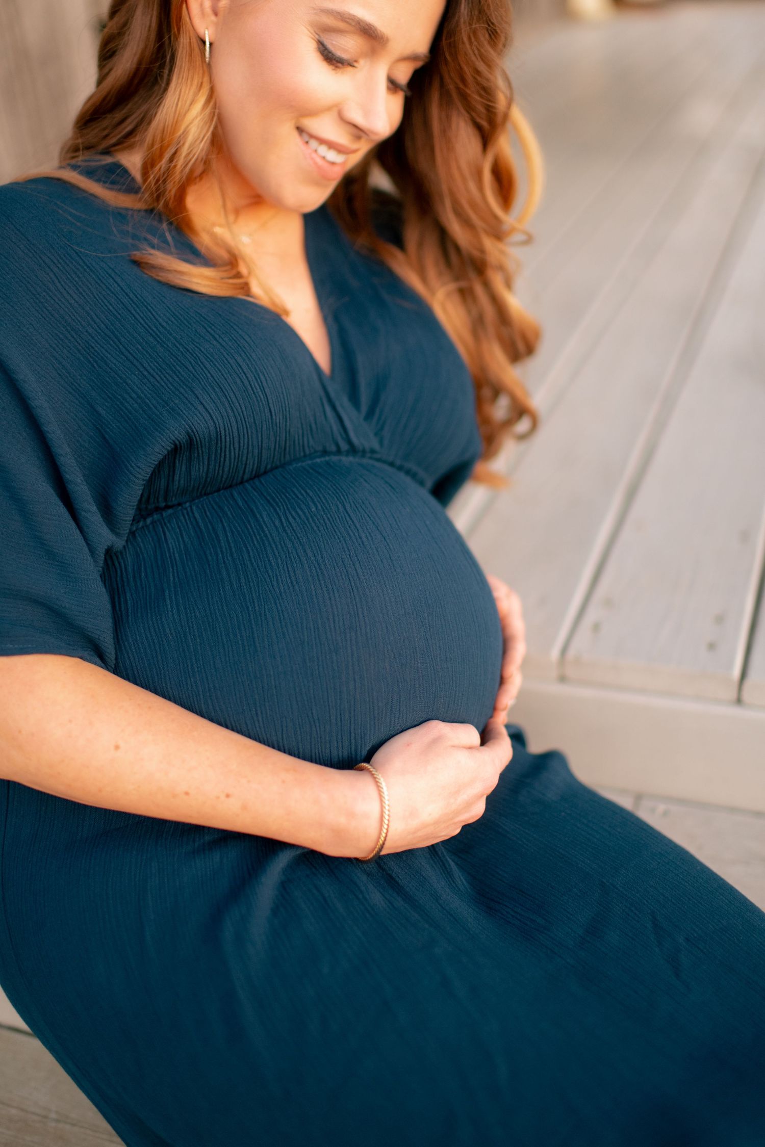 Pregnant woman in navy blue dress gently cradles her baby bump while smiling warmly.