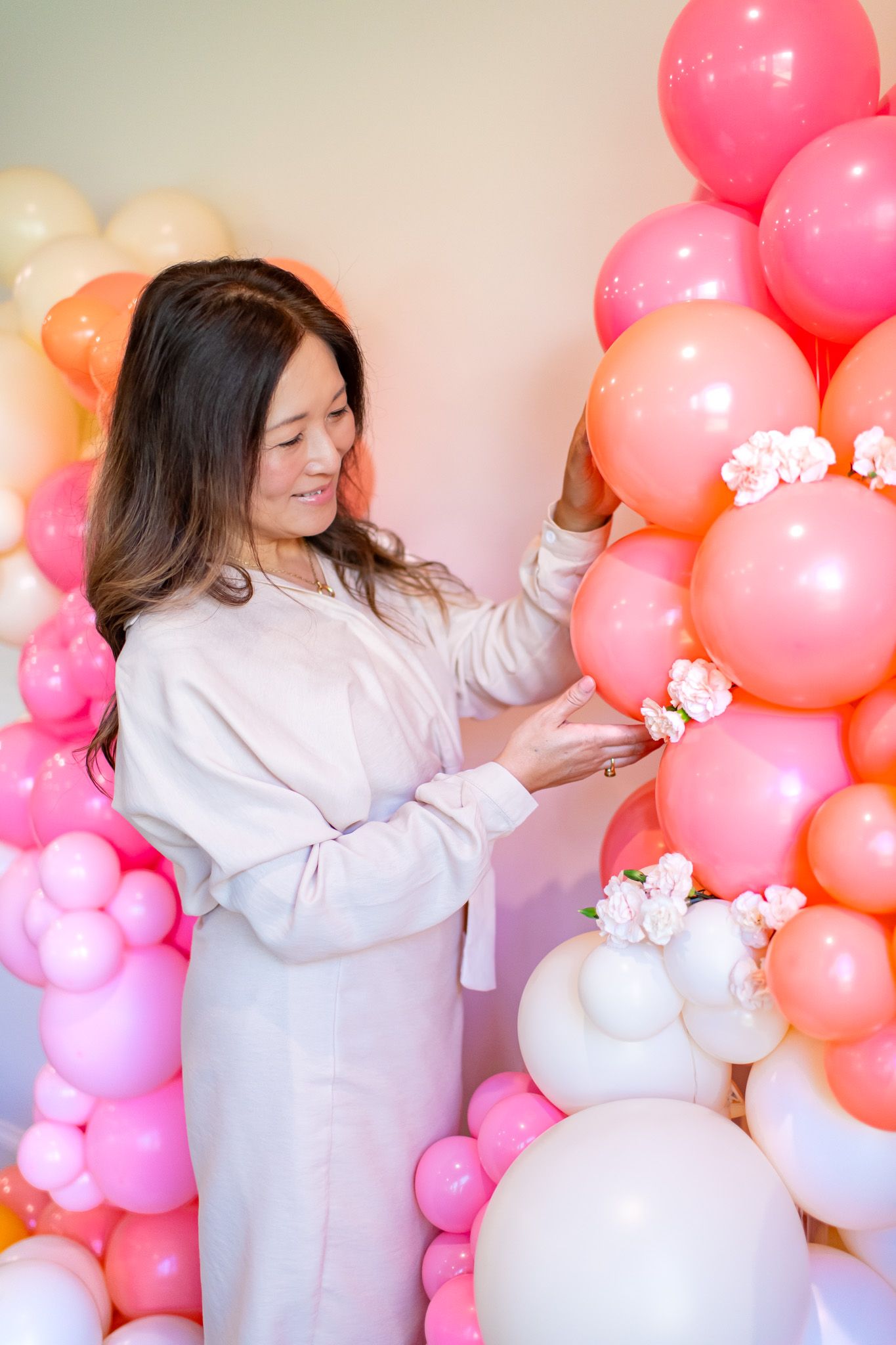 Woman in white dress interacts with balloon wall installation featuring pink, coral and white balloons.