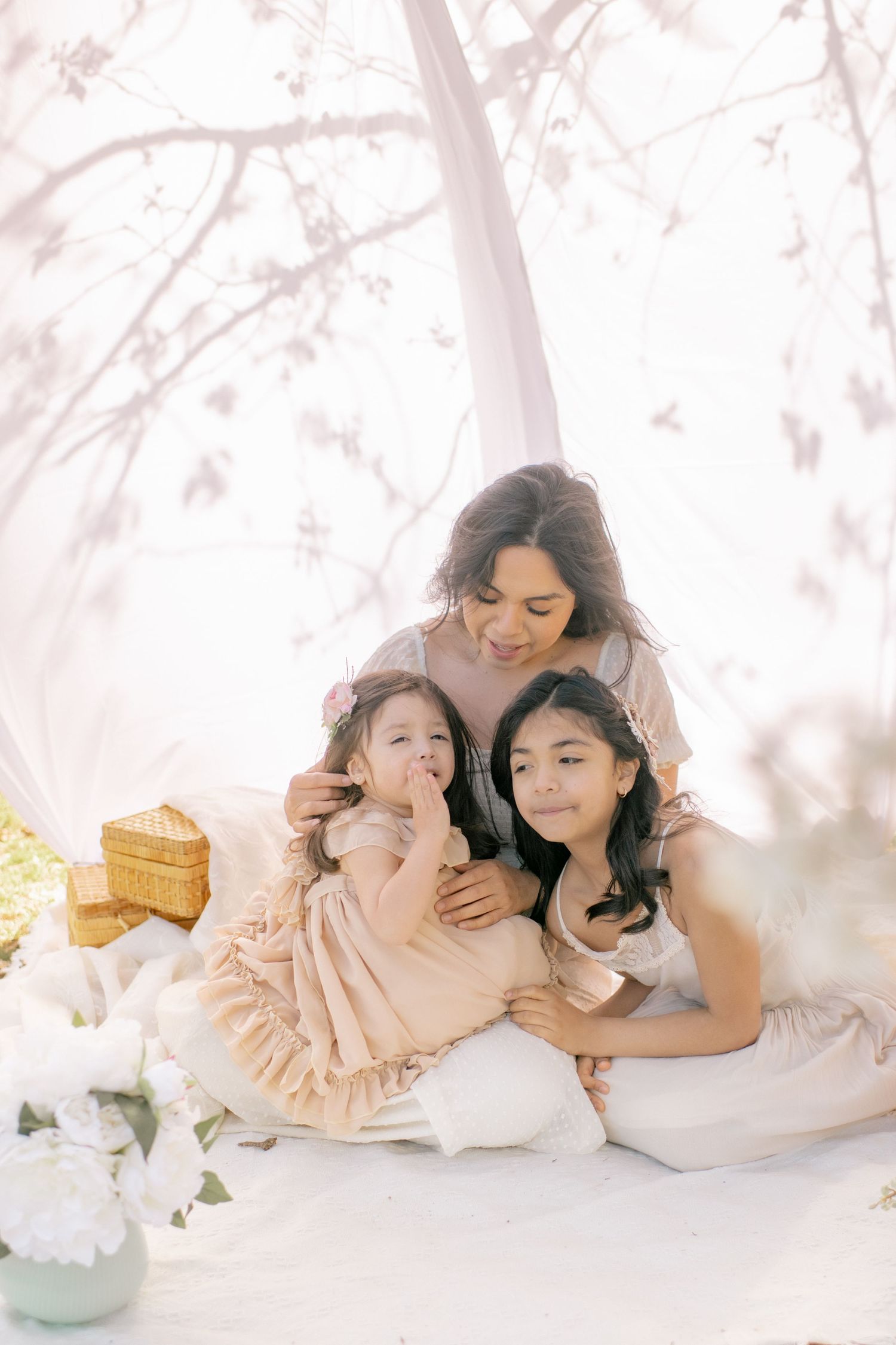 A tender spring portrait of three people in white dresses sitting together amidst soft pink cherry blossoms.