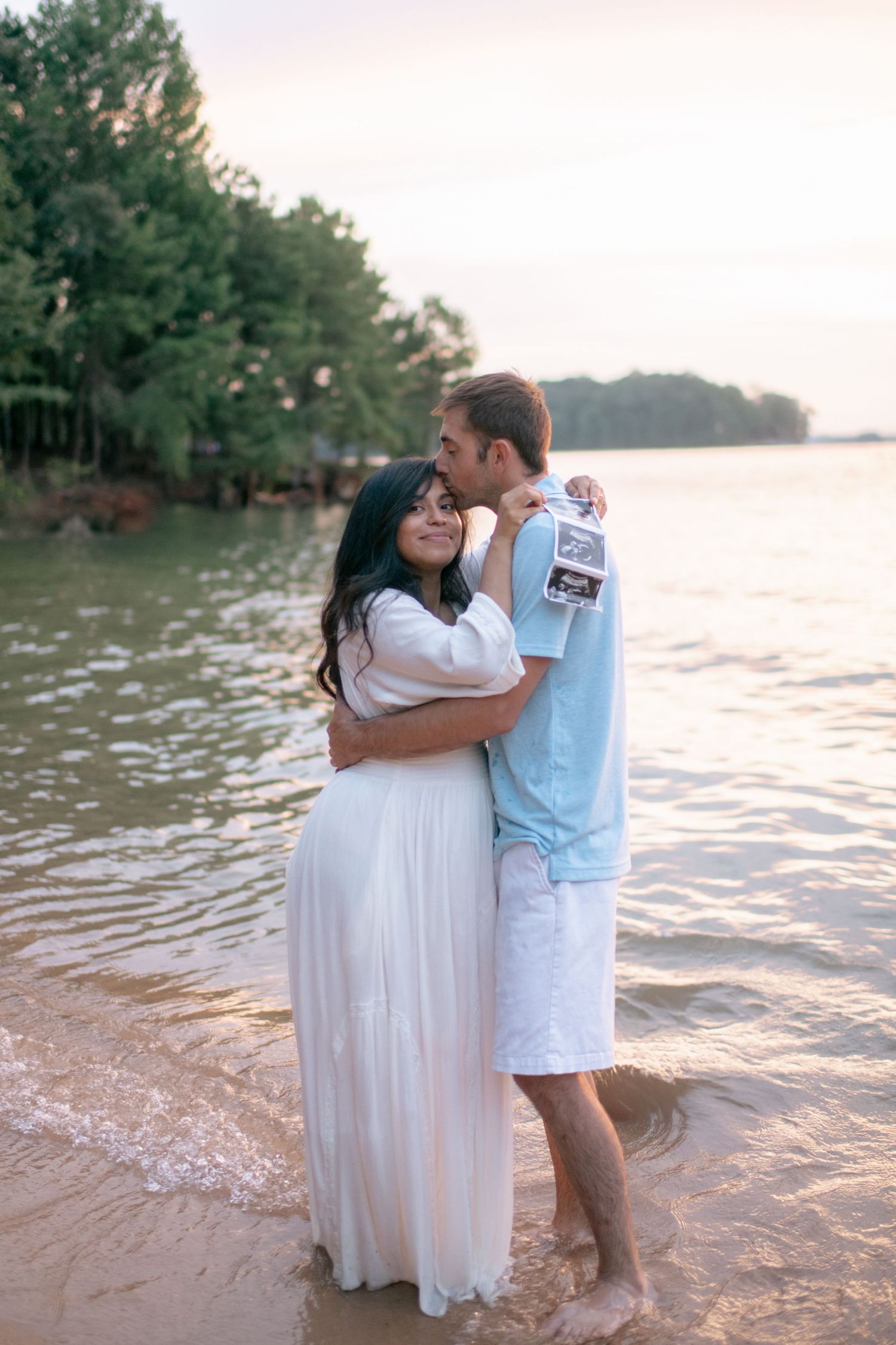 Couple embraces on sandy beach shoreline during sunset with lake and pine forest in background.