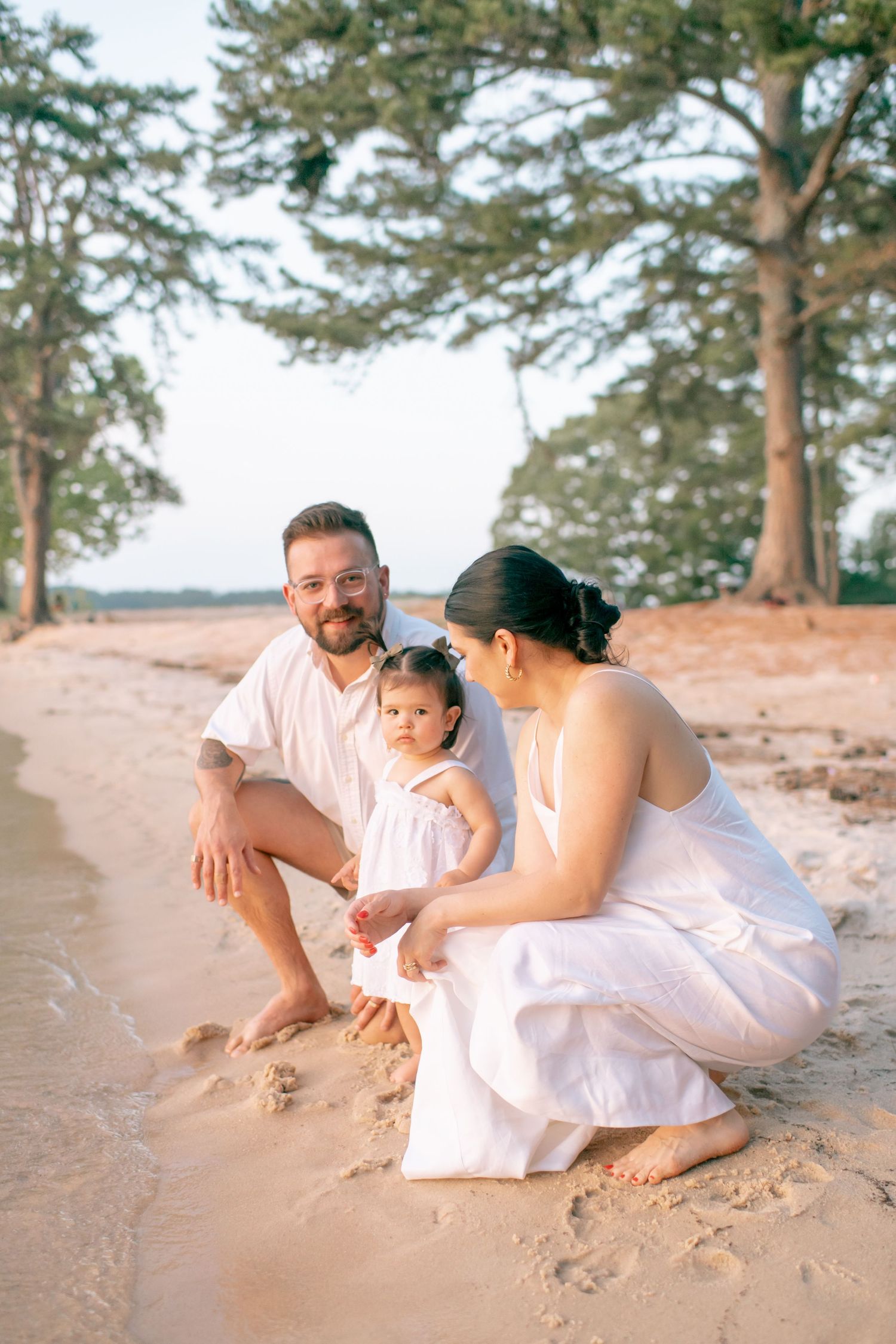 Family relaxes together on a sandy beach with pine trees in the background.