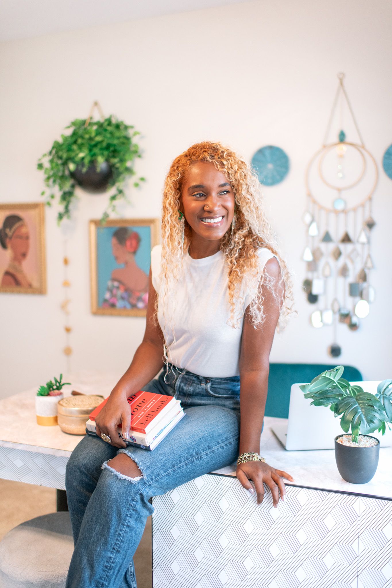 Person in casual attire sits on white surface with decorative wall hangings and plants in background.