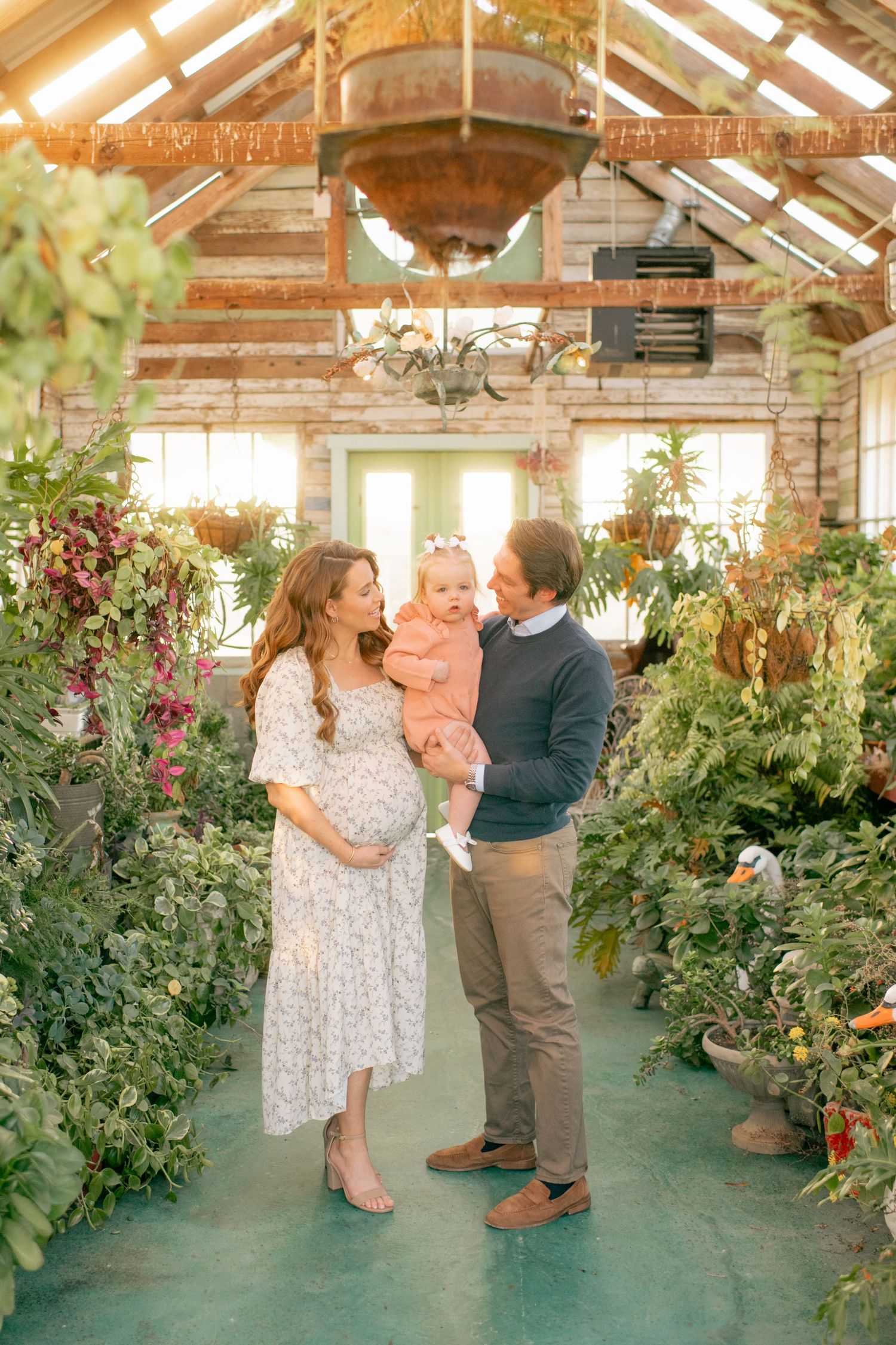 Family standing together in a sunlit greenhouse filled with lush plants and flowers in a warm, ethereal setting.